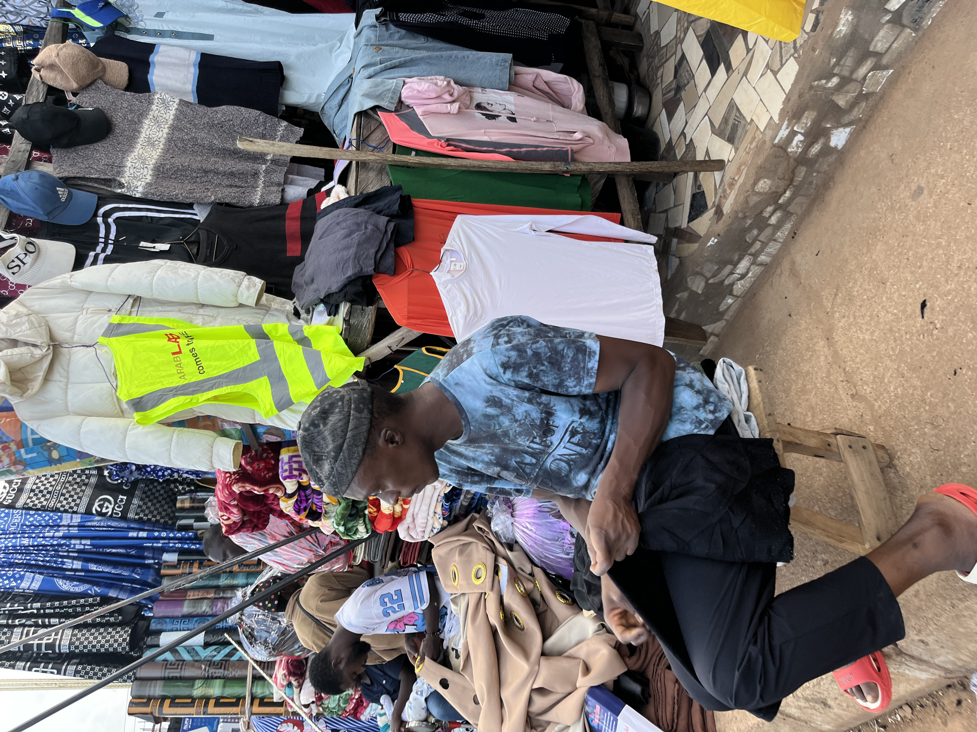 The image captures a bustling market scene in Bukuru, Nigeria, where various clothing items are displayed for sale. The primary subjects are two men, likely vendors or shoppers, interacting amidst the merchandise. One man, wearing a blue tie-dye t-shirt and a black cap, is seen holding a pair of black trousers. He appears focused on the transaction or the items he is handling. Another man is partially visible in the foreground, bent over and possibly assisting with the goods.
The setting is an outdoor market with a rustic wooden stall structure holding stacks of folded clothes. These include shirts, jackets, trousers, and what appear to be blankets or throws, featuring a variety of colors and patterns. Notable objects include a bright yellow reflective vest with text, likely for safety, draped over a white jacket. Another section of the stall displays rolled fabrics with intricate patterns, some bearing logos that resemble "GUCCI."
The overall impression is one of commercial activity and everyday life in a Nigerian market. The ground is a mix of dirt and scattered paving stones, indicating an unpaved or partially paved area. The lighting suggests it is daytime, possibly late morning or early afternoon, with the sun casting some shadows, though the weather appears fair. There are no overt emotional expressions visible, but the focused demeanor of the individuals suggests a typical market interaction. The presence of diverse clothing items and the general atmosphere point to a vibrant local marketplace.