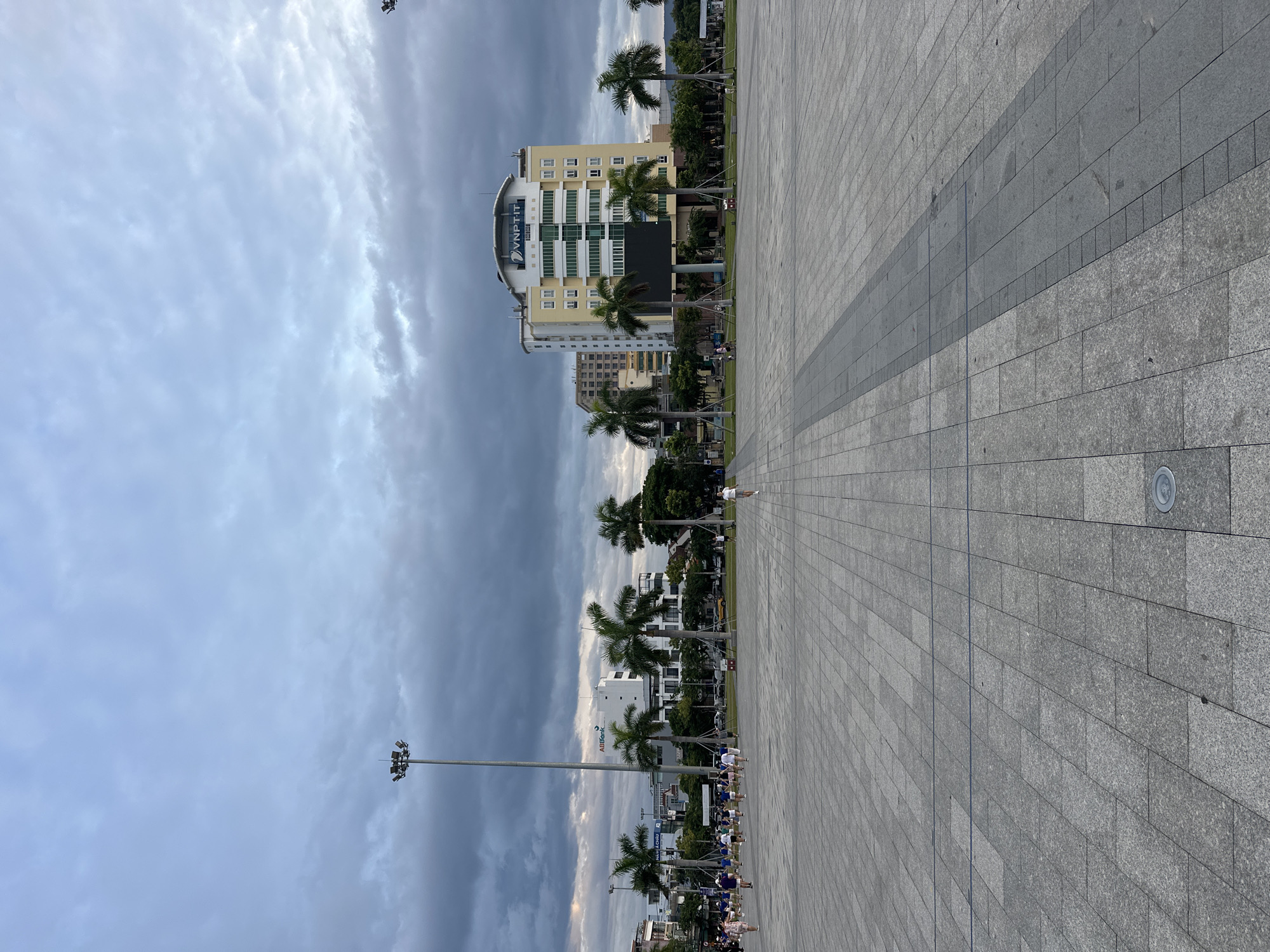 This image captures a wide view of a paved plaza in Đà Nẵng, Vietnam, likely the Quảng trường 2 tháng 9 (September 2nd Square). The sky is overcast with dramatic, dark clouds, suggesting an impending storm or the aftermath of one. Tall lampposts stand sentinel on the left side of the plaza, with their lights off, illuminated by the ambient daylight.
In the middle ground, a group of people, possibly a sports team given their matching blue and white attire, are gathered and walking across the plaza. Further back, several palm trees stand tall, interspersed with modern buildings. The most prominent building is a multi-story yellow structure with the "VNPT-IT" logo visible at the top. Adjacent to it is a white building with the "ABBANK" logo.
The foreground is dominated by the expansive, tiled surface of the plaza, with a distinct pattern of lighter and darker grey rectangular tiles. A few scattered individuals are walking across this open space. The overall impression is one of a large urban public space under a moody sky, with signs of activity and modern infrastructure.