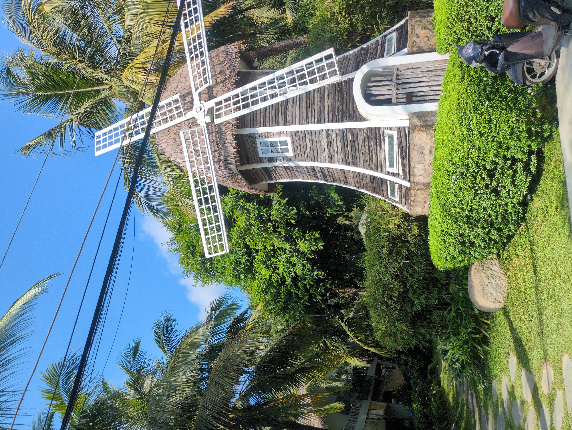 The image shows a thatched-roof windmill in Phú Quốc, Vietnam. The windmill has four white blades, a dark wood exterior with white trim, and a small window. A decorative archway is visible on one side. The windmill is surrounded by lush green vegetation, including palm trees and hedges. A motorcycle is parked nearby. Power lines run across the blue sky in the background. The environment suggests a tropical or subtropical climate.