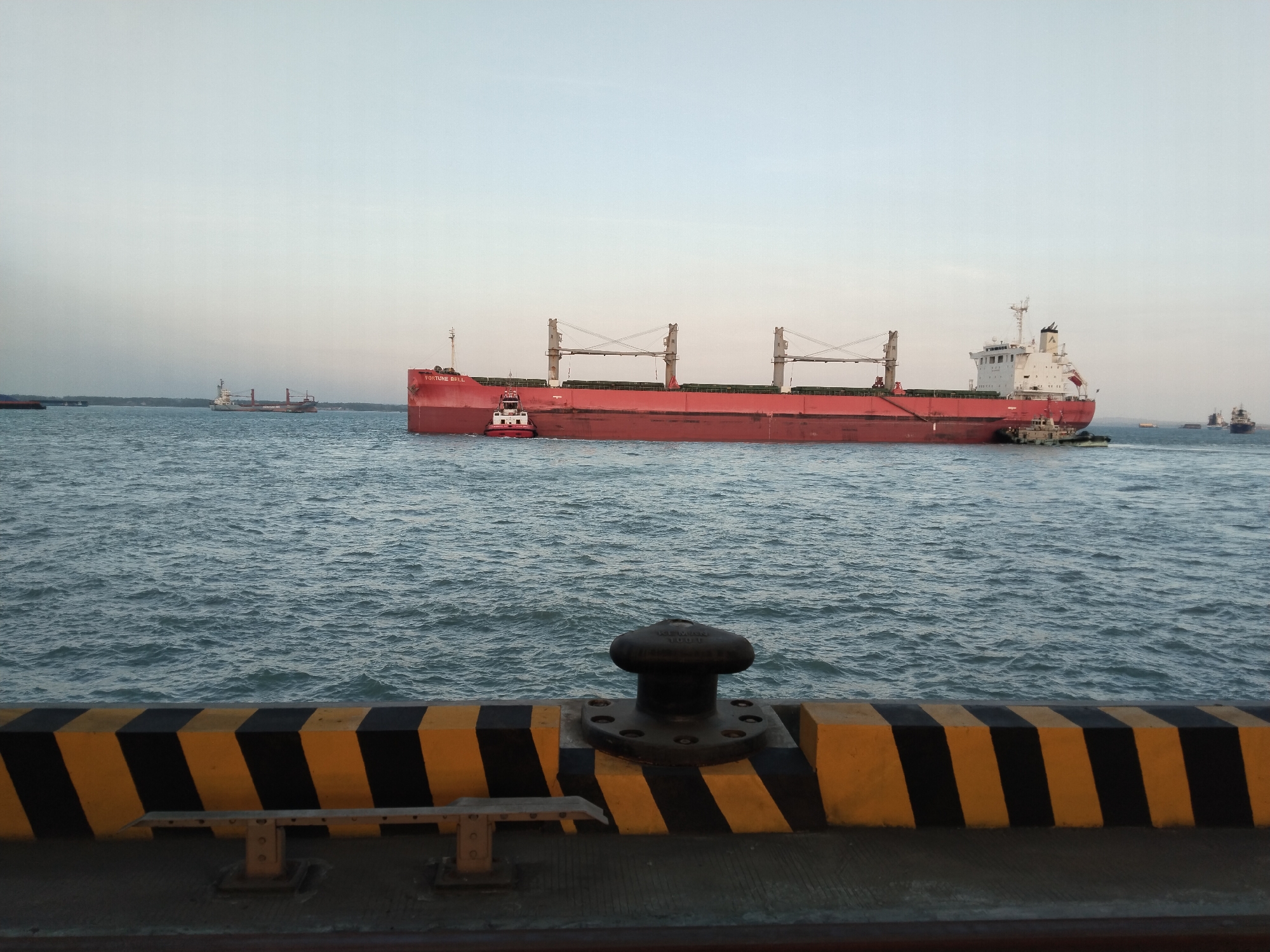 A large red cargo ship, named "FORTUNE BELL", is being assisted by a small tugboat in the waters off Pernajuh, Indonesia. The cargo ship is a bulk carrier with cranes visible on its deck. The tugboat is positioned to the port side of the cargo ship, appearing to guide it. In the foreground, a dock edge with yellow and black striped safety markings is visible, along with a metal bollard and a bench. Other smaller vessels are present in the distance, and the sky is clear and overcast. The water is choppy.