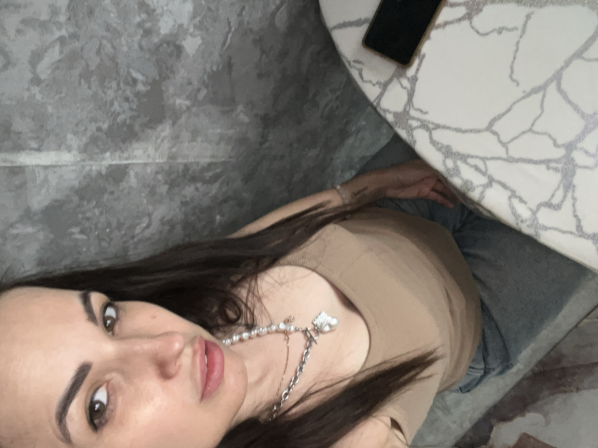 The image captures an overhead, angled view of a woman in an indoor setting.
**Subjects:** The primary subject is a woman with long, dark, straight hair, brown eyes, and distinct dark eyebrows. She is wearing a light brown or tan-colored top, possibly a tank top or camisole, and blue jeans. Around her neck, she wears a two-part necklace featuring pearls on one strand and a silver chain with a small leafy charm on the other. Her right forearm bears a faint tattoo. Next to her, on a table, a dark-colored smartphone with a golden or metallic edge is visible.
**Scene and Setting:** The woman is seated or leaning next to a table with a white top patterned with grey or silver veining, resembling marble or quartz. The background consists of a mottled grey wall or textured surface. The overall setting appears to be an indoor space with modern decor.
**Activity or Event:** The woman is looking directly at the camera with slightly parted lips, suggesting she is posing for a photograph, likely a selfie given the close-up and high-angle perspective.
**Notable Details:** The photograph is taken from an elevated viewpoint and is rotated, creating a dynamic and somewhat disorienting composition. The lighting is soft and even, typical of indoor artificial light or diffuse natural light, without strong shadows indicating a specific time of day. Her expression is calm and direct. The tattoo on her right forearm is a single word in cursive, which is difficult to decipher clearly but appears to be "Charmant" or something similar.
**Visible Text:** A faint cursive word tattooed on the woman's right forearm.