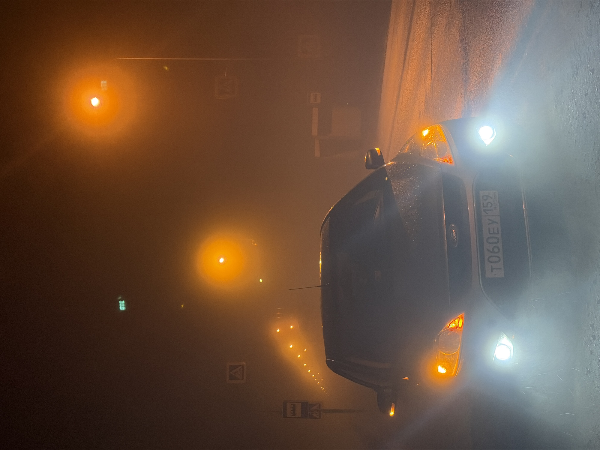 A dark gray Ford Focus car with headlights on is parked on the side of a road in heavy fog. The license plate reads "T060EY 159 RUS". Streetlights illuminate the fog in a warm, orange hue. Road signs are visible in the fog, indicating this is likely an urban area. Based on the license plate number, this is in Noviy, Russia. There are no people visible. The environment appears to be cold and damp.
