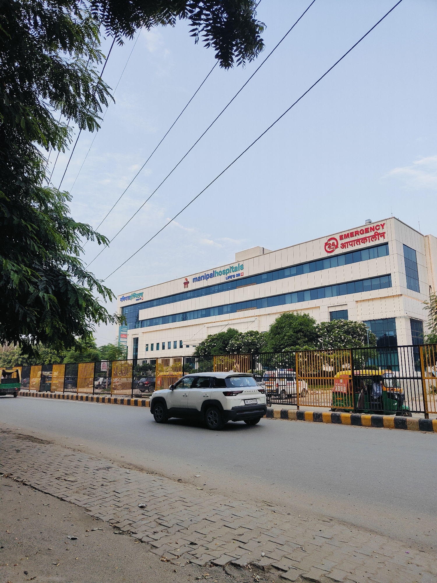 The image captures a street scene in Gurgaon, India, featuring the Manipal Hospitals building. The hospital, a modern white structure with blue-tinted windows, has prominent signage in English and Hindi, including "Manipalhospitals" and "EMERGENCY आपातकालीन". A white SUV is driving on the road in front of the hospital, passing a black and yellow striped barrier and a fence. Beyond the fence, several trees and other vehicles, including an auto-rickshaw, are visible. The sky is a light blue with some clouds, and power lines crisscross the upper portion of the image. The street in the foreground is paved with grey asphalt and has a tiled sidewalk on the left. The overall impression is a typical urban environment during the daytime.