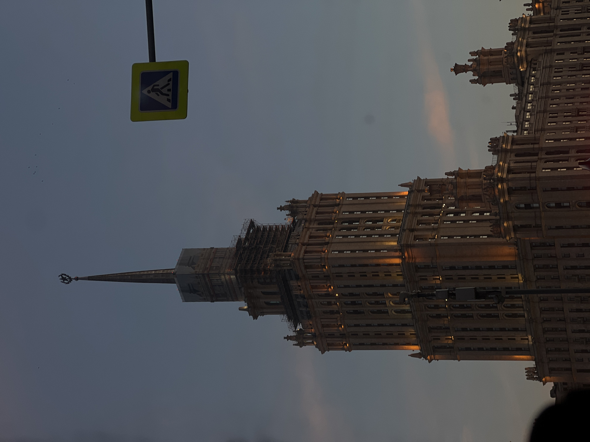 The image displays the Hotel Ukraina (Radisson Collection Hotel, Moscow) in Moscow, Russia, at twilight. The multi-tiered, ornate Stalinist Empire style building is illuminated with warm yellow light from numerous windows, highlighting its architectural features. Scaffolding is visible around the upper sections of the central tower and the base of its prominent spire, which is topped with a star emblem. The sky is a deep blue-grey, with subtle pink clouds visible in the background. In the upper right quadrant, a yellow square pedestrian crossing sign, featuring a black border and a white walking figure on a dark blue background, is mounted on a dark pole. A dark, vertical utility pole partially obscures the lower-mid section of the building. Several small, dark avian shapes are visible against the sky in the upper left. An indistinct dark form occupies the extreme lower foreground, suggesting a partial view of a person or object.