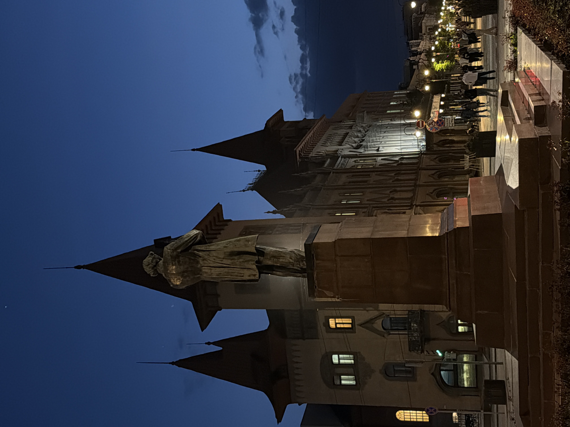 At night in Saratov, Russia, a statue stands in front of a building with spires. People stroll along a sidewalk illuminated by streetlights. The sky is a deep blue, with clouds visible above the building.