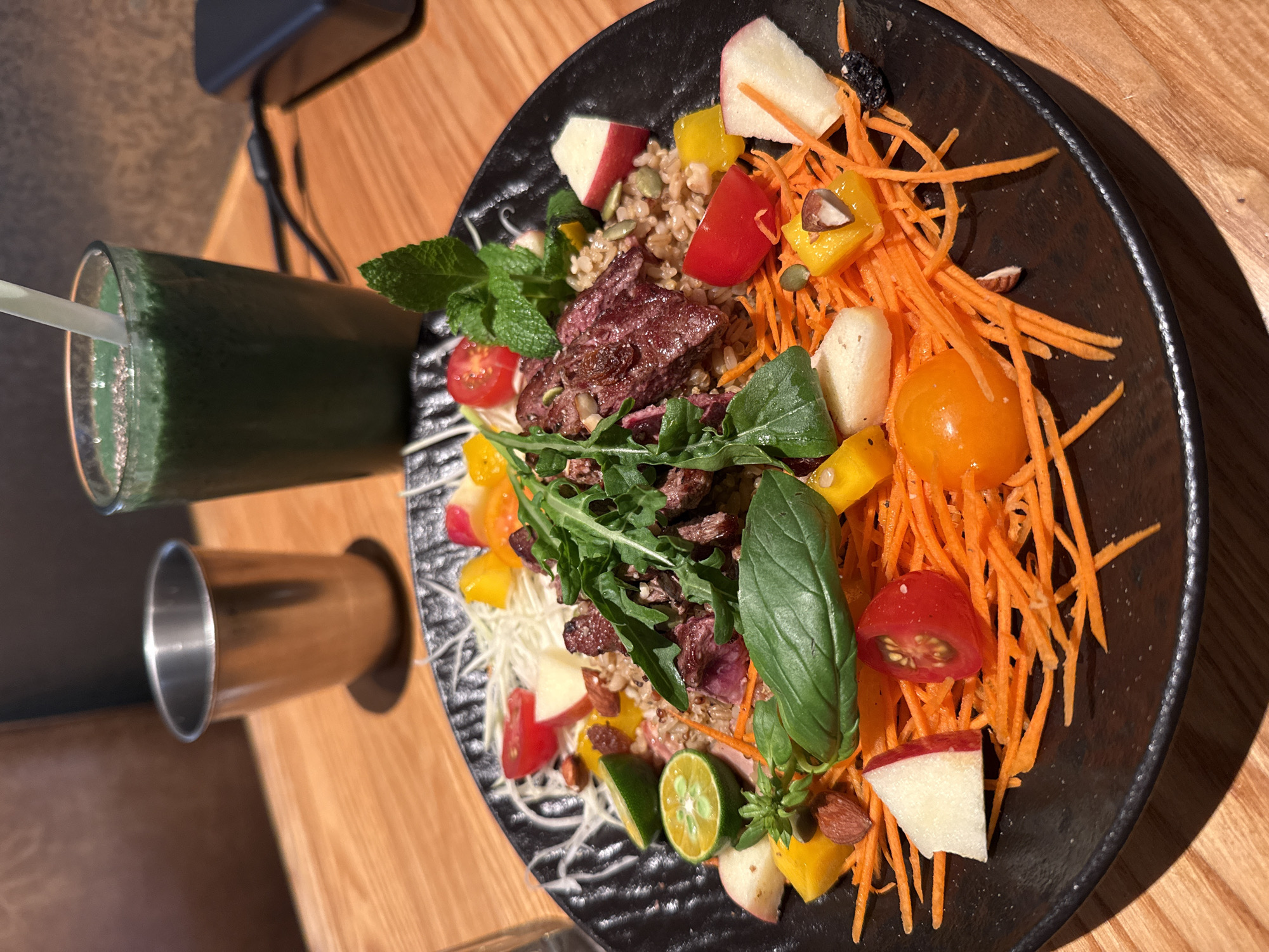 A high-angle shot shows a plate of salad and a beverage on a wooden table. The salad, served on a black plate, contains shredded carrots, pieces of red and yellow tomatoes, apple slices, grain, greens (likely arugula and basil), what appears to be cooked meat, and herbs like mint. A green beverage in a glass with a straw stands beside the plate, and a metal cup is visible near the glass. The scene is lit by natural light, creating shadows on the right side of the plate and the table. There's no indication of the location being in Yuzhong District, China, as there are no recognizable landmarks or text in the image.