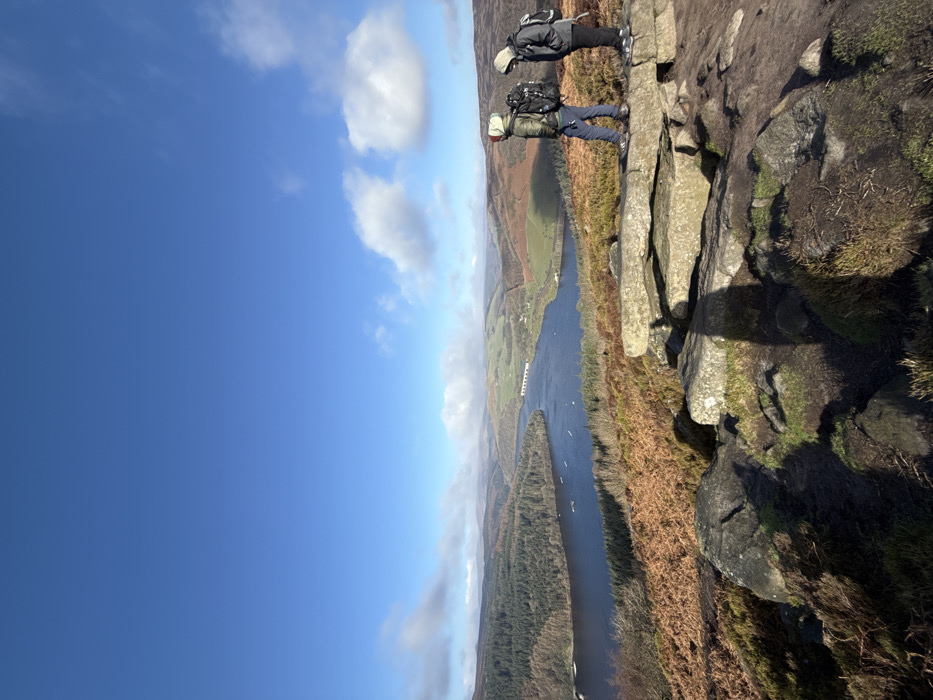 The image captures an expansive outdoor scene on a clear, bright day under a blue sky with scattered white clouds, located in the Peak District, High Peak, United Kingdom, overlooking Ladybower Reservoir. In the foreground, two individuals, dressed in hiking attire and carrying backpacks, stand on a flat, light-grey rock outcrop amidst brown heather and green moss. The individual on the left wears a khaki jacket, light blue trousers, and a light-colored hood, while the individual on the right wears a dark hooded jacket and dark trousers. Both are facing right, observing the distant landscape. Below them, the dark blue waters of Ladybower Reservoir stretch across the mid-ground, containing several small boats and spanned by a distant grey arched viaduct. The reservoir is flanked by dense green coniferous forest on the left bank and rolling hills with mixed brown and green vegetation on the right. Further rolling hills extend into the background beneath the horizon.