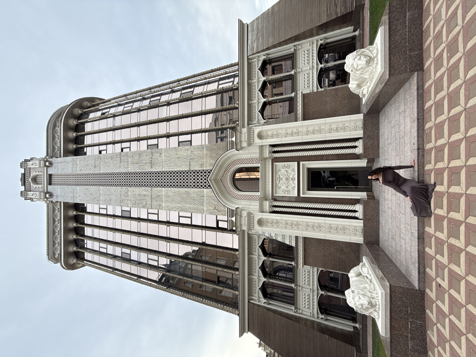 A low-angle, wide shot captures a woman in a long, dark grey dress sitting on the steps of a large, ornate building. The building, identified as the "Millennium Tower" in Kazan, Russia, is characterized by its distinctive architecture, featuring a central, tall tower with decorative patterns and a facade adorned with columns and intricate stonework. Two white lion statues flank the entrance. The woman is positioned on the steps, leaning forward with her right hand supporting her on the step, facing left with her head turned towards the camera. The foreground shows a patterned stone pathway. The sky is overcast, and reflections of buildings are visible on the mirrored windows of the tower.