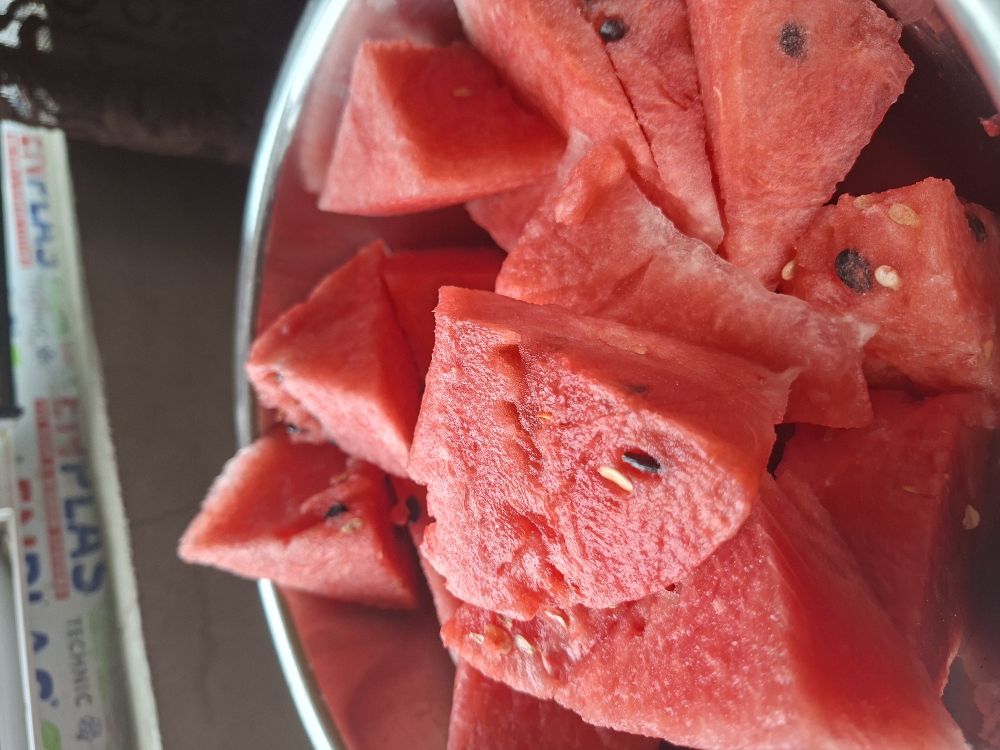The image shows a close-up of a metal bowl filled with cut pieces of watermelon. The watermelon slices are triangular and a vibrant red color, with some visible black seeds and lighter colored seeds. The texture of the watermelon flesh is visible, appearing juicy and slightly fibrous. The bowl is partially visible around the edges, indicating it is a shallow, wide bowl. In the background, out of focus, there is a surface with some text that appears to be part of a label or packaging, with the words "TECHNIC" and "EMPLAS" partially visible. Above this, a blurred object or surface is present, possibly a shelf or window sill. The lighting suggests an indoor setting. There are no people visible in the image. The image does not provide any information or context to determine its location in the city of Tbilisi, Georgia.