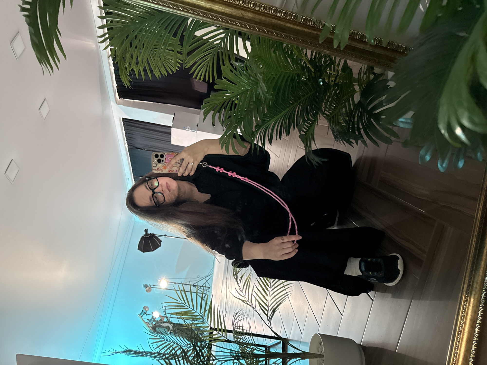 A woman is visible in the foreground, likely taking a selfie in a mirror. She has long brown hair and is wearing glasses. She is dressed in black and has a pink beaded lanyard around her neck. In her hand, she is holding a mobile phone.
The background is a room with mixed elements. There are plants visible, both behind the woman in the mirror and in front. One plant is in a white pot. A soft blue light emanates from the wall behind the plants, which may be a decorative lighting fixture. The floor is made of wooden planks.
While it is not possible to confirm the location with complete certainty based solely on the visual information, the provided context suggests the city of Chaikovsky, Russia as the location.