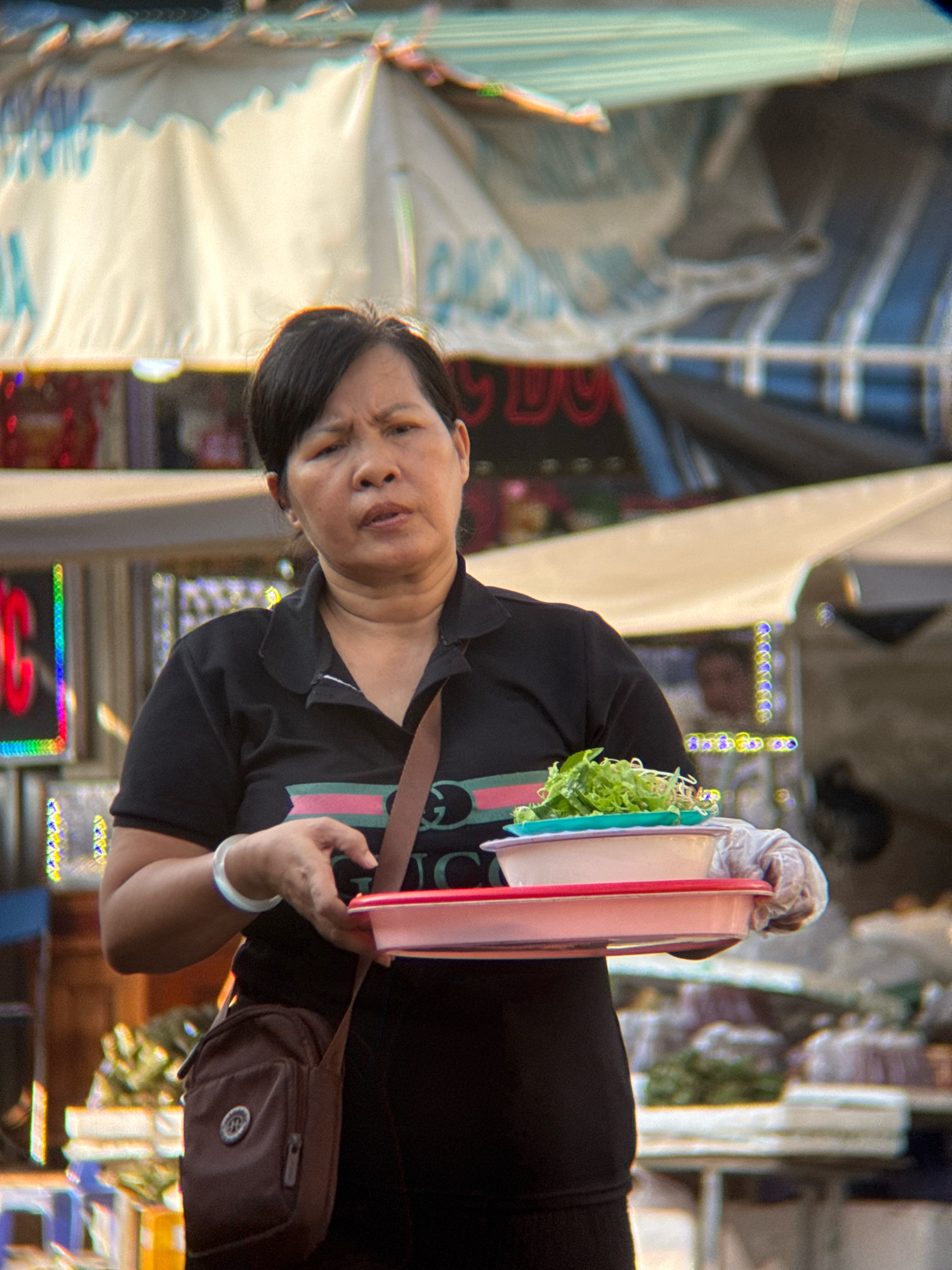 A middle-aged Asian woman with short black hair, wearing a black polo shirt with a green and pink stripe across the chest, is standing in what appears to be an outdoor market stall in Nha Trang, Vietnam. She is holding a stack of plastic food containers, the top one containing a portion of fresh green leafy vegetables and bean sprouts, which she is presenting to the viewer. Her facial expression is neutral. The background is a blurred market scene with awnings, stalls, and some colorful lights. She is wearing a white bracelet on her left wrist and has a dark brown crossbody bag slung over her right shoulder. Her hands are covered with white disposable gloves.