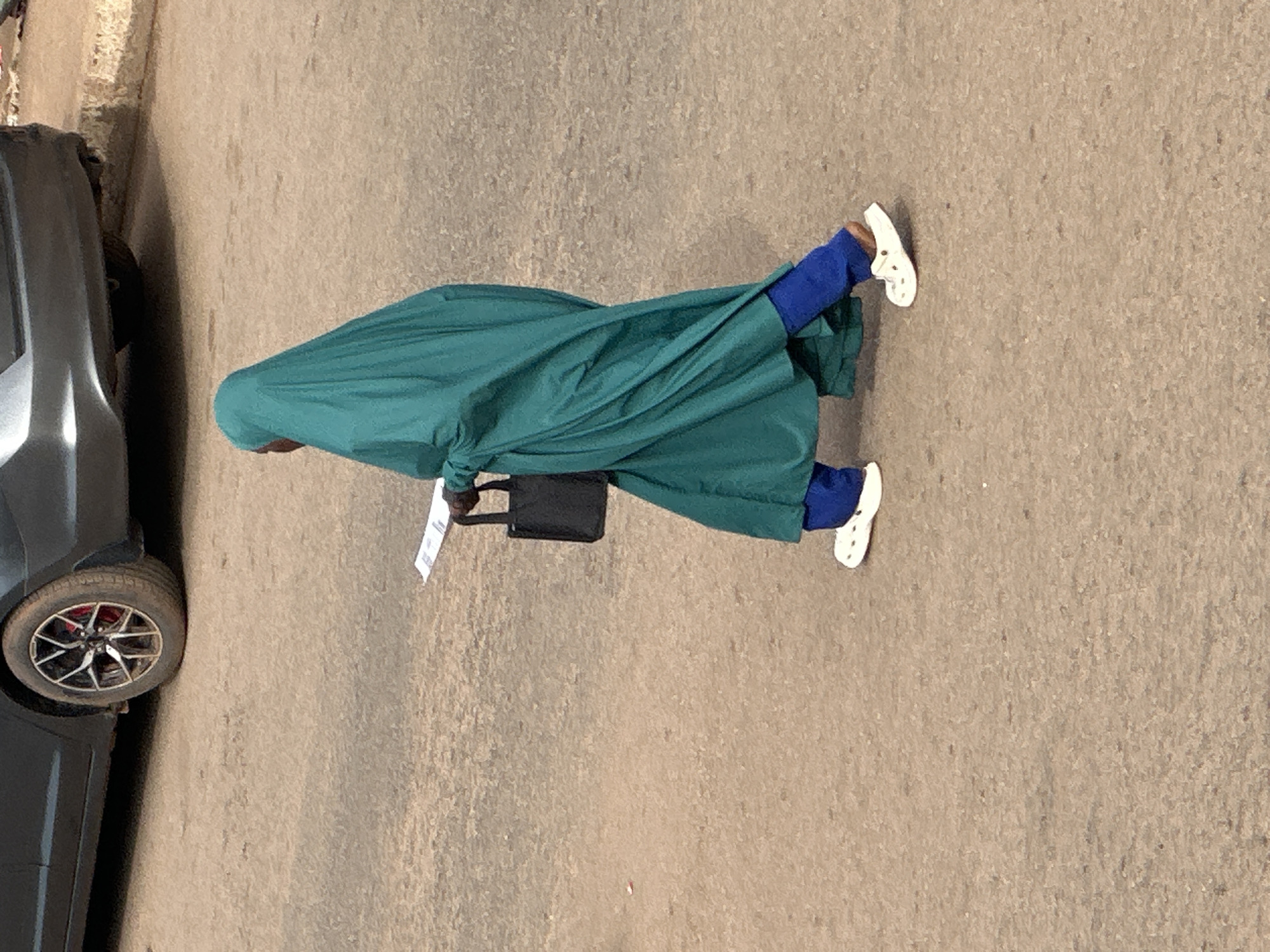 A person wearing a teal abaya and blue trousers walks on a dirt road in Bukuru, Nigeria. The person is also wearing white Crocs and carrying a black tote bag. A dark-colored car is partially visible in the background.