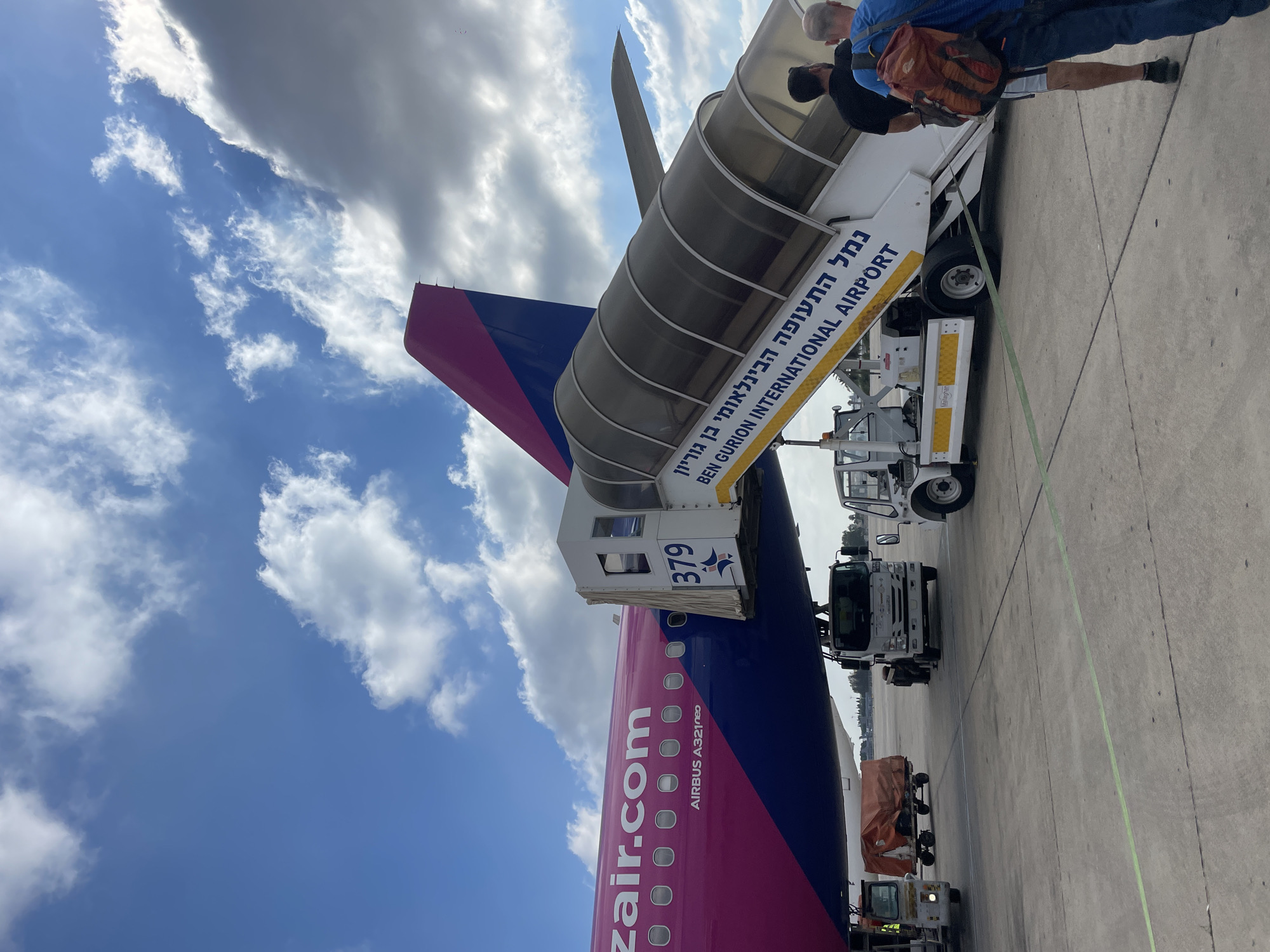 A pink and blue Wizz Air Airbus A321neo airplane is parked on an airport tarmac under a cloudy blue sky. A mobile passenger staircase is connected to the aircraft's door, with the words "BEN GURION INTERNATIONAL AIRPORT" printed on its side in English and Hebrew. Two men, one with a red backpack, are walking up the stairs, presumably boarding the plane. Several ground vehicles, including a baggage tug and a tow truck, are visible near the aircraft. The scene appears to be at Ben Gurion International Airport near Tel Aviv, Israel, not the city of Yagel.