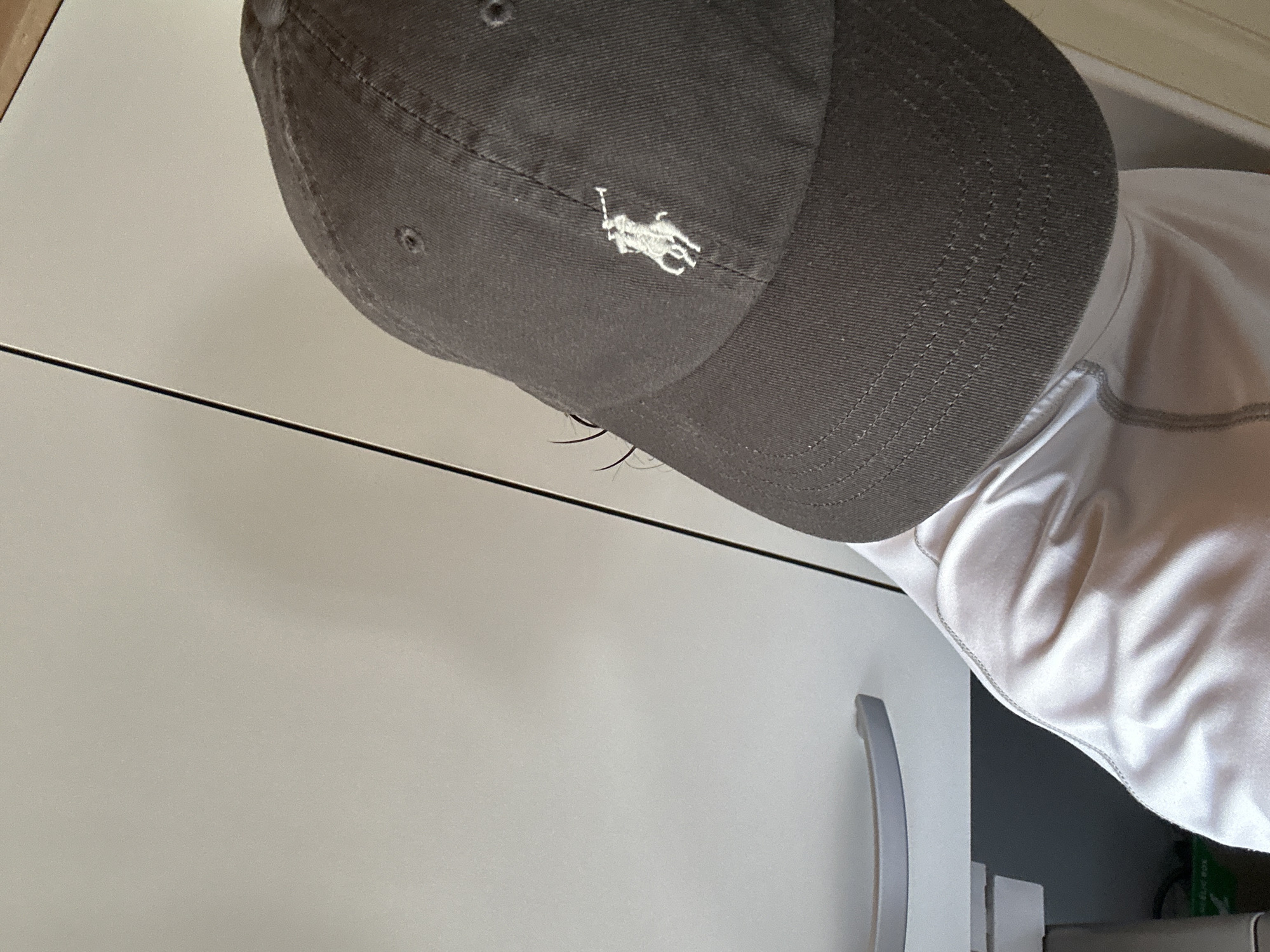 The image shows a close-up, rotated view of a person wearing a dark grey baseball cap with a white embroidered logo of a polo player on a horse. The cap appears to be made of fabric, with visible stitching around the brim and a vent hole on the crown. The person is also wearing a light-colored, possibly white, t-shirt with grey stitching along the seams. The background is a plain, light-colored surface, possibly a wall or cabinet, with a horizontal black line running across it. To the right, a portion of a silver handle is visible on what appears to be a piece of furniture, perhaps a cabinet or drawer. The overall lighting suggests an indoor setting, possibly during the daytime, as there are no harsh shadows. The image is focused on the cap and upper torso of the person. There is no visible text in the image.