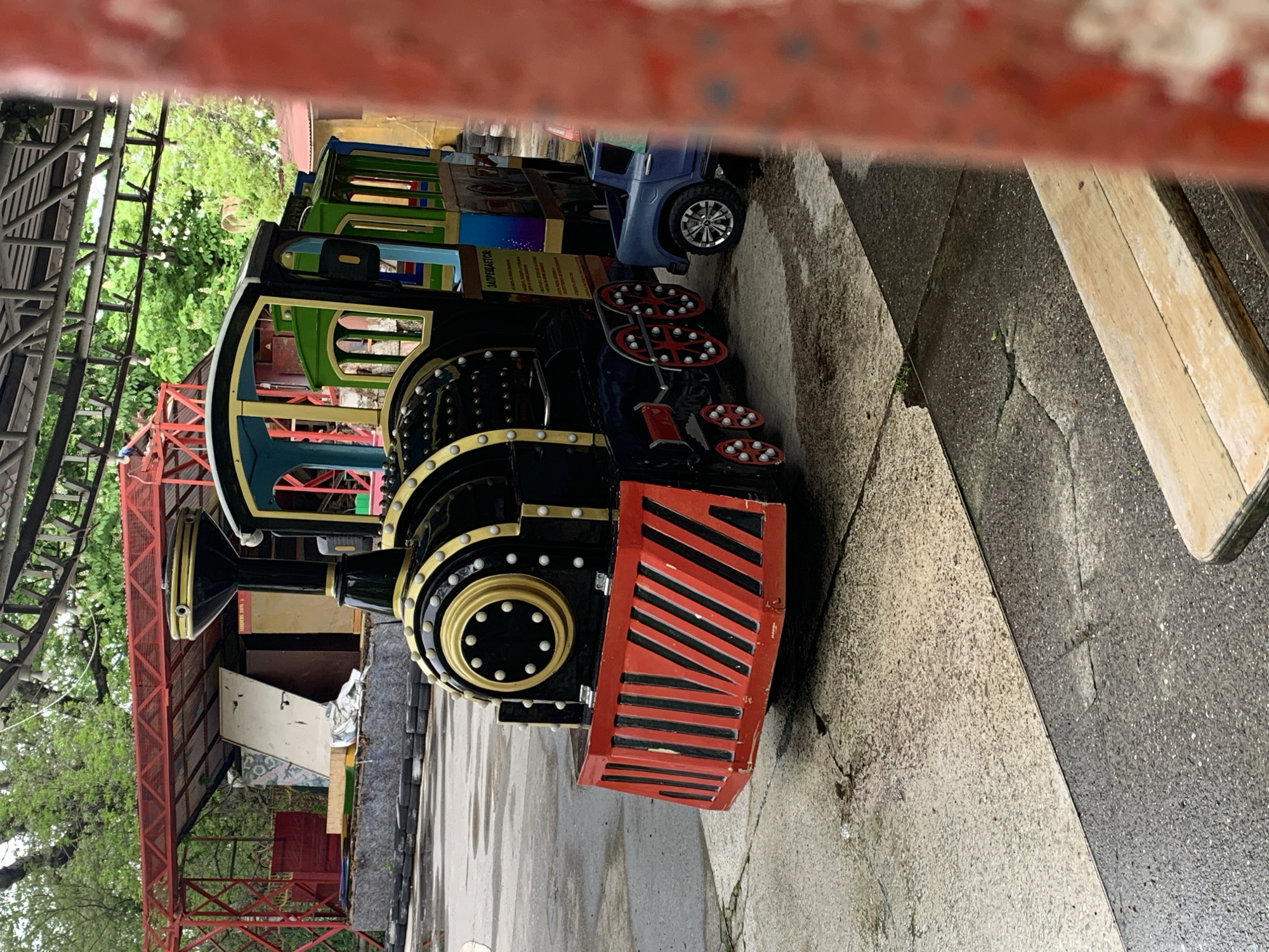 A partially obstructed view of a small amusement park train is captured, located outdoors in Stavropol, Russia. The train, painted in black, red, green, and blue, is positioned on a concrete surface. The front of the train is most visible, with a red striped bumper and gold trim. Other cars of the train, in green and blue, are behind the engine. A portion of a red metal structure is visible above and to the side of the train. The background includes some foliage and a glimpse of a building. The image is taken from a low angle.