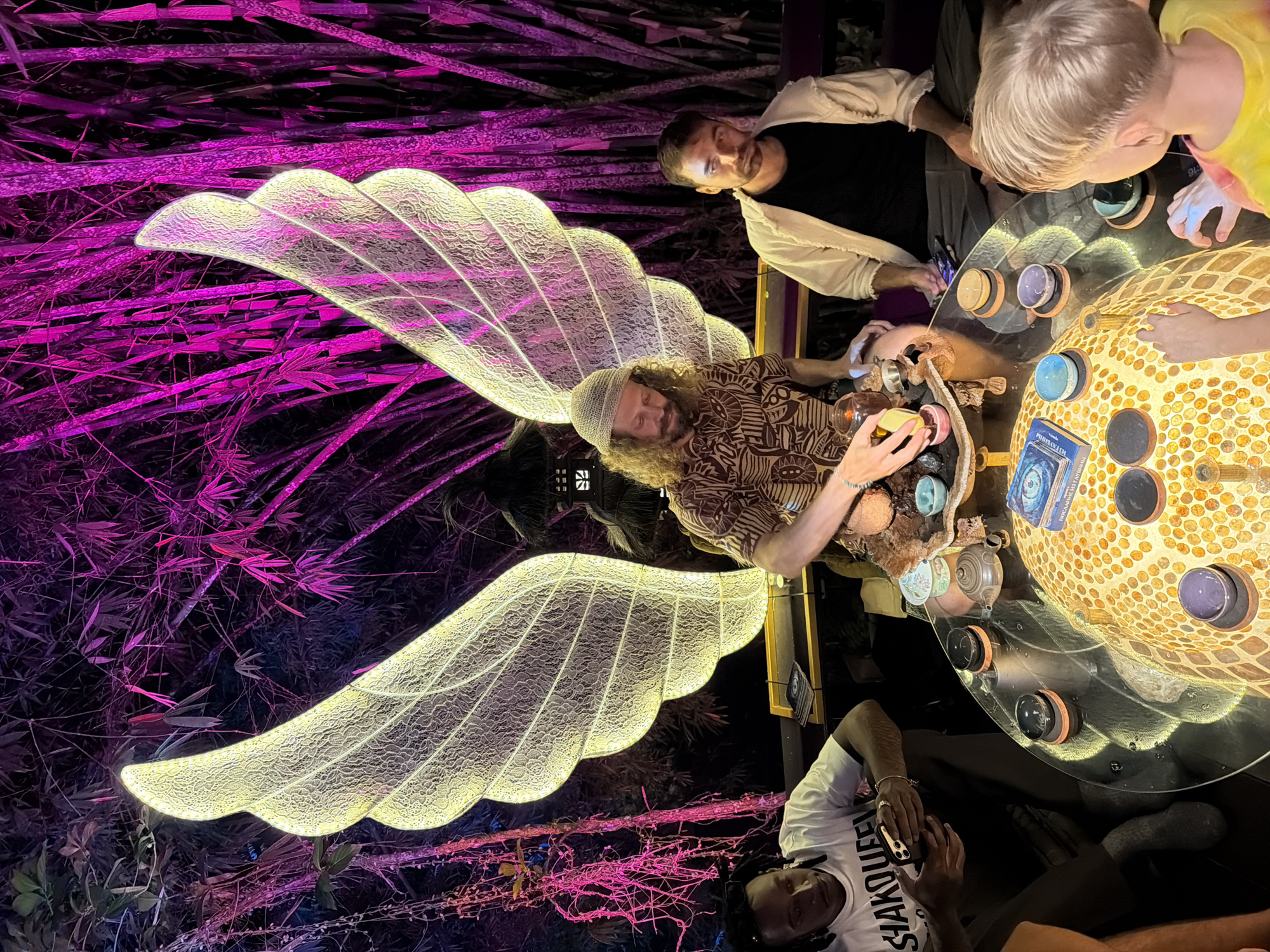 The image depicts a group of people gathered around a circular table in an outdoor setting, illuminated by a large, illuminated wing-like structure and pink lighting on bamboo stalks. In the foreground, a young child with blonde hair is looking at the table. To the left, a Black man in a white t-shirt is holding a smartphone. In the center, a man with curly blonde hair and a patterned shirt is interacting with items on the table, which include small bowls, a teapot, and what appear to be tarot cards or oracle cards. To the right of the center man, another man in a cream-colored jacket over a black shirt is seated. The table itself is glass-topped and features a mosaic-like illuminated base. The backdrop consists of dark vegetation, primarily bamboo, with vibrant pink lighting. The overall atmosphere suggests a spiritual or meditative gathering. The location is identified as Blahbatuh, Indonesia.
