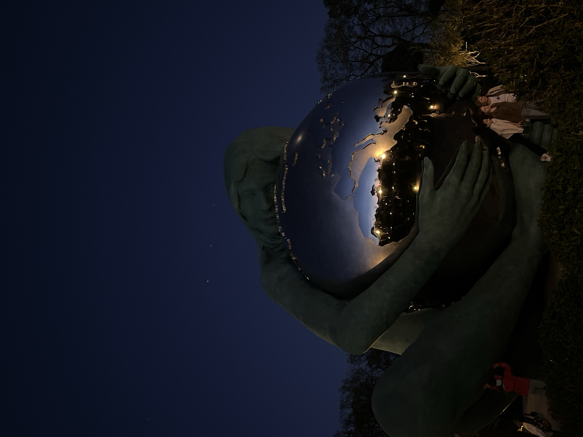 The image captures a large, dark statue at night in Krasnodar, Russia. The statue depicts a figure, likely female, with a serene expression, embracing a highly reflective spherical object that resembles the Earth. The figure's left arm is prominently visible, and her hands are wrapped around the sphere as if holding it protectively.
The spherical object, a stylized globe, gleams with reflections of artificial lights, highlighting continental shapes that appear to be Europe and Africa. The surface of the globe acts as a mirror, clearly reflecting three individuals lying on their backs on the ground, positioned within the statue's embrace. They are casually dressed, with one person in a light top and dark pants, another in a brown jacket, and a third in a lighter shirt. A fourth person in a red jacket is faintly visible near the bottom right, by the statue's leg.
The background is a deep navy blue night sky, with a few faint stars visible in the upper left portion of the frame. To the right and behind the statue, dark trees and bushes are silhouetted against the dim light, indicating an outdoor, park-like setting. The overall lighting suggests it is late evening or night. There is no visible text in the image.