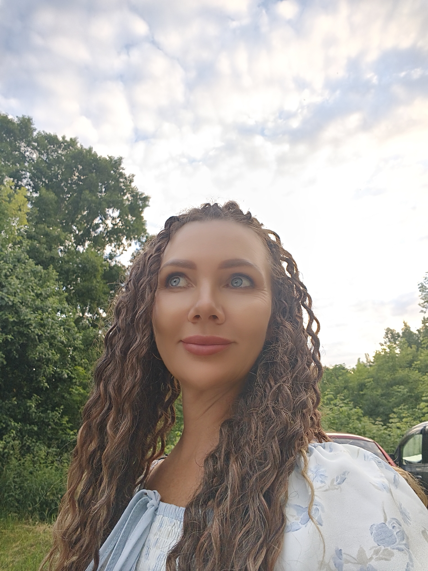 A medium shot captures a woman with long, curly brown hair styled in braids, looking upwards and to the left. She has fair skin, blue eyes, and is wearing a light blue and white floral top. The background consists of lush green trees and a cloudy sky, suggesting an outdoor setting. A portion of a red car and a dark vehicle are visible on the right side of the image. The scene is set in the city of Chaikovsky, Russia.