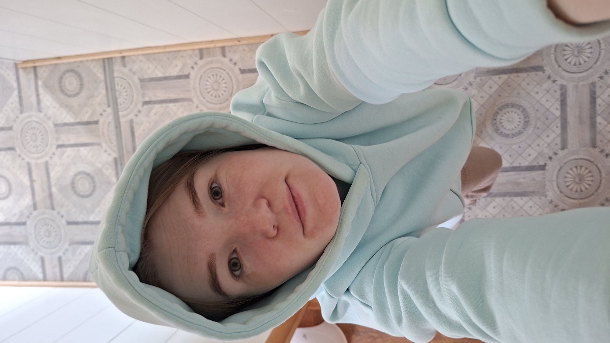 A high-angle selfie captures a young person indoors, looking up towards the camera with a gentle expression. The subject, likely female, has fair skin with noticeable freckles across her nose and cheeks, light brown hair, and green eyes. She wears a light blue or mint-colored hooded sweatshirt, with the hood pulled up around her head, framing her face. Her lips are slightly parted in a subtle, pleasant smile.
The camera is positioned directly above her, looking down, and the image is oriented with her head towards the left side of the frame. Her arms, clad in the light blue hoodie, extend towards the right side.
The background primarily consists of distinct patterned flooring, featuring circular and geometric designs in shades of grey and white. Above the patterned floor, a light-colored wall, possibly shiplap or paneled, is visible, with a thin wooden trim running horizontally across it. The lighting appears bright and even, suggesting it is daytime indoors, possibly illuminated by natural light or well-distributed artificial lighting. There is no visible text in the image. The setting appears to be a residential or domestic space, consistent with the location context of Большой Букор, Russia.