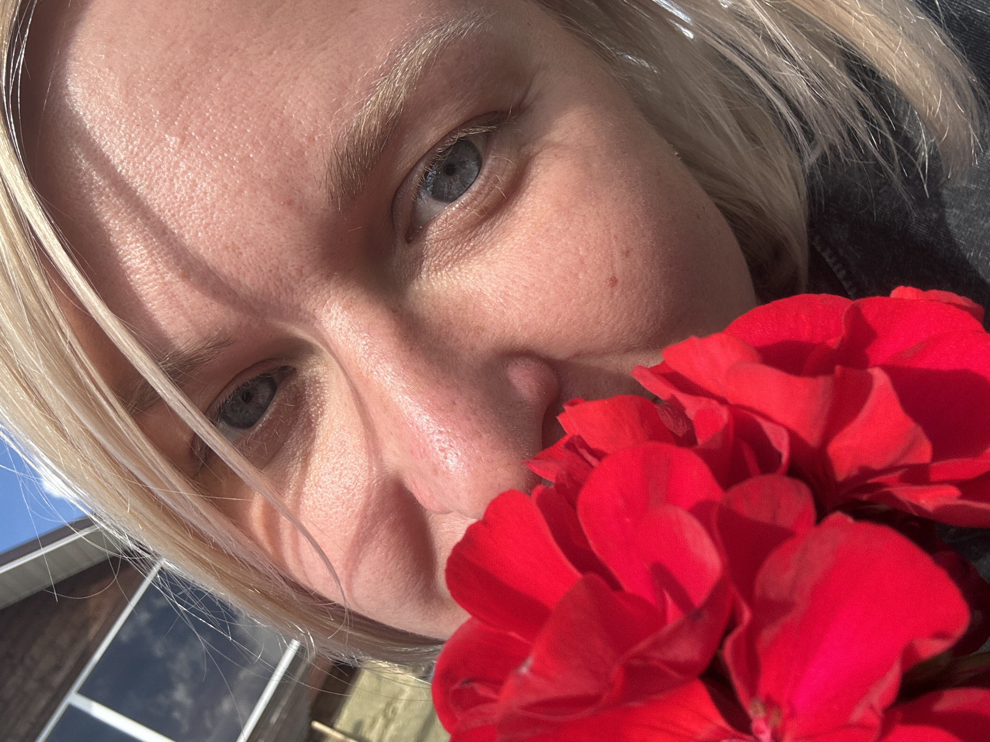 The media file is a close-up, low-angle selfie of a person with blonde hair. The subject is holding bright red flowers, which partially obscure their face. Their blue eyes are visible, and the sunlight casts strong shadows across their forehead and cheek. The background is slightly out of focus, showing a building with windows that reflect the sky, suggesting an outdoor setting. The sky appears blue with some white clouds, indicating it is daytime. The overall impression is a candid moment, possibly a personal snapshot taken outdoors on a sunny day. No visible text is present in the image. The location context provided is Chaikovsky, Russia.