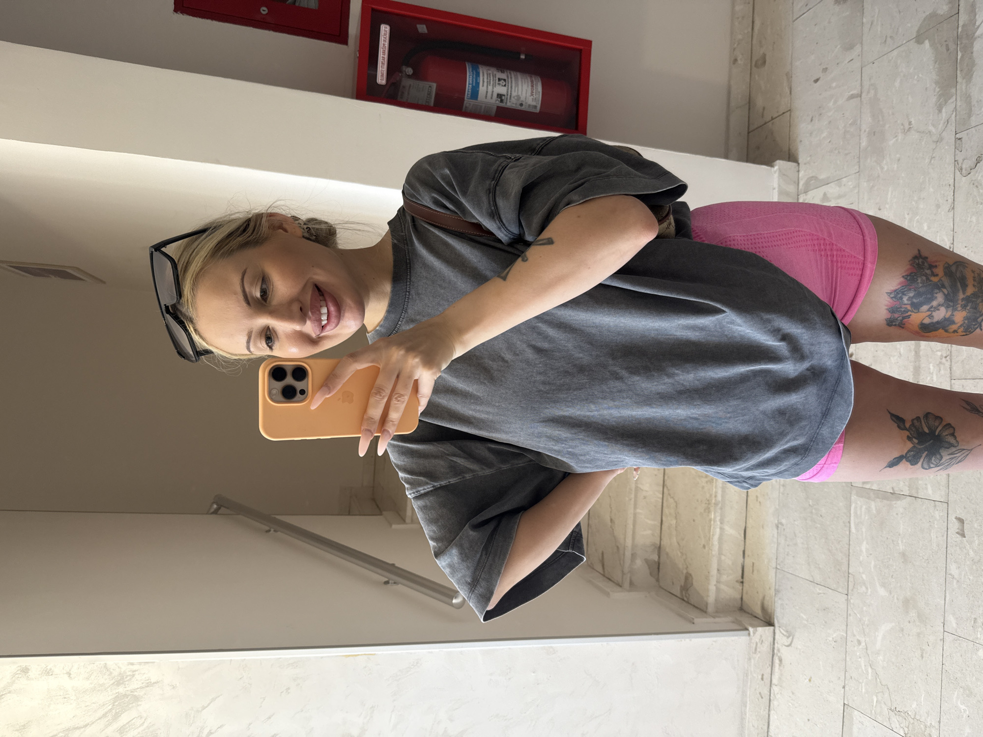 In an indoor setting, a young woman with blonde hair pulled back and sunglasses perched on her head takes a selfie in a mirror. She is wearing an oversized gray t-shirt and bright pink shorts. Tattoos are visible on her legs, including a floral design and a depiction of two people kissing. She is holding a bright orange smartphone, which is capturing the reflection. Behind her, a staircase with a metal railing is visible, and to her right, a red fire extinguisher is mounted on the wall. The floor is tiled with a light-colored marble pattern.