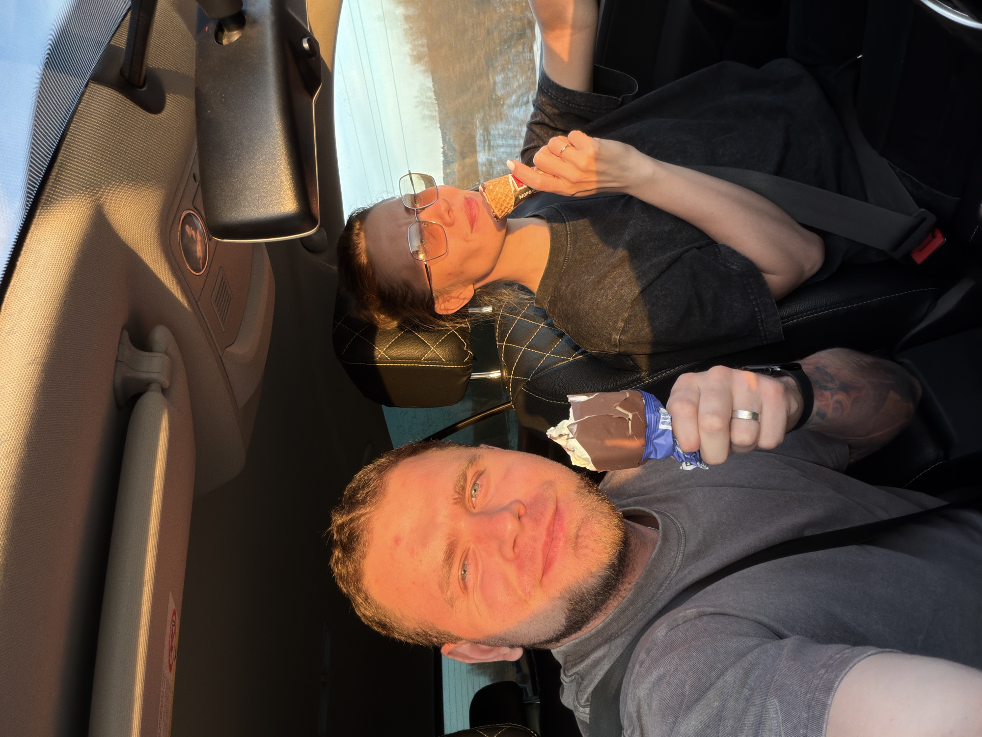 Two people are in a car, presumably traveling. The person in the foreground is male with blue eyes and a beard, wearing a gray shirt and holding a partially eaten chocolate-covered ice cream bar. He is wearing a seatbelt and has tattoos on his arm. Behind him, a female with glasses is leaning back, also wearing a seatbelt, and eating an ice cream cone. The car's interior is visible, including the rearview mirror and sun visor. The location, Полозово, Russia, cannot be confirmed or denied by visual elements in the provided image.