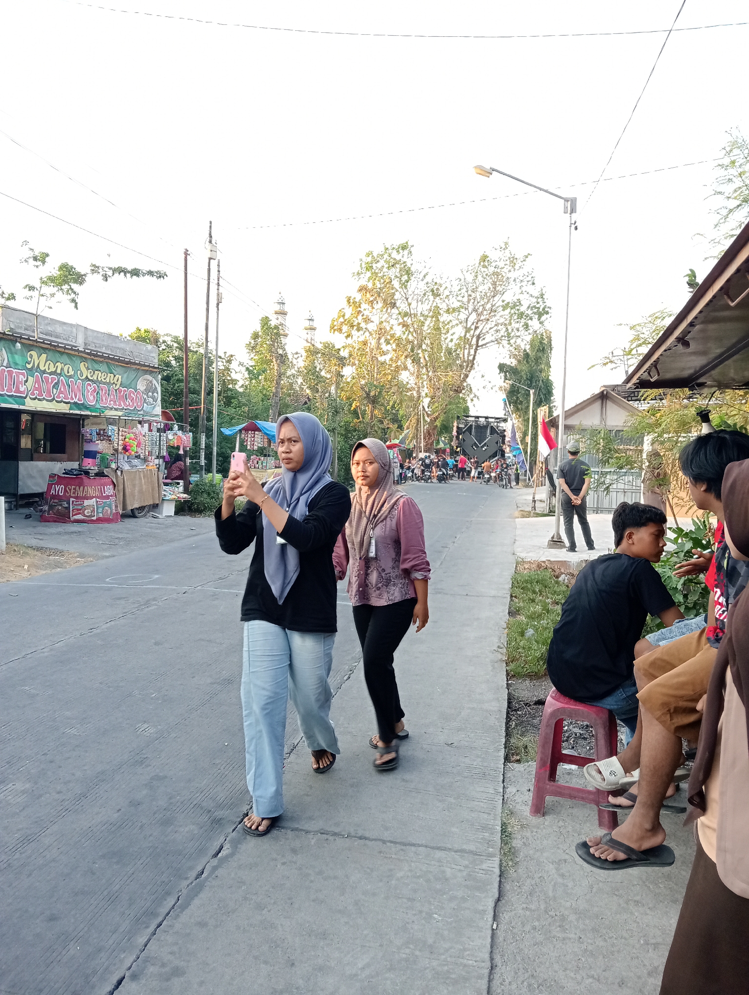 The image depicts a street scene in Tanjunganyar, Indonesia, during what appears to be late afternoon or early evening. The sky is bright and overcast, suggesting daylight. On the left side of the image, a roadside eatery with a sign reading "Moro Seneng NIE AYAM & BAKSO" is visible, along with various displays of goods. A woman wearing a light blue hijab and a black shirt is standing on the asphalt road, holding a pink smartphone and appearing to take a photo. To her right, another woman in a mauve hijab and patterned shirt walks past.
Further down the road, a gathering of people and motorcycles is visible, with a large sound system or stage setup in the background. A man in a grey cap and dark pants stands facing away from the camera near a lamppost with an Indonesian flag attached. On the right side of the road, a group of young men are seated on a low red stool and on the ground. One man wears a black t-shirt and denim shorts, another a black t-shirt, and others are partially visible. They appear to be engaged in conversation. The environment includes a paved road, a concrete sidewalk, and vegetation along the roadside. The overall atmosphere suggests a community gathering or event.