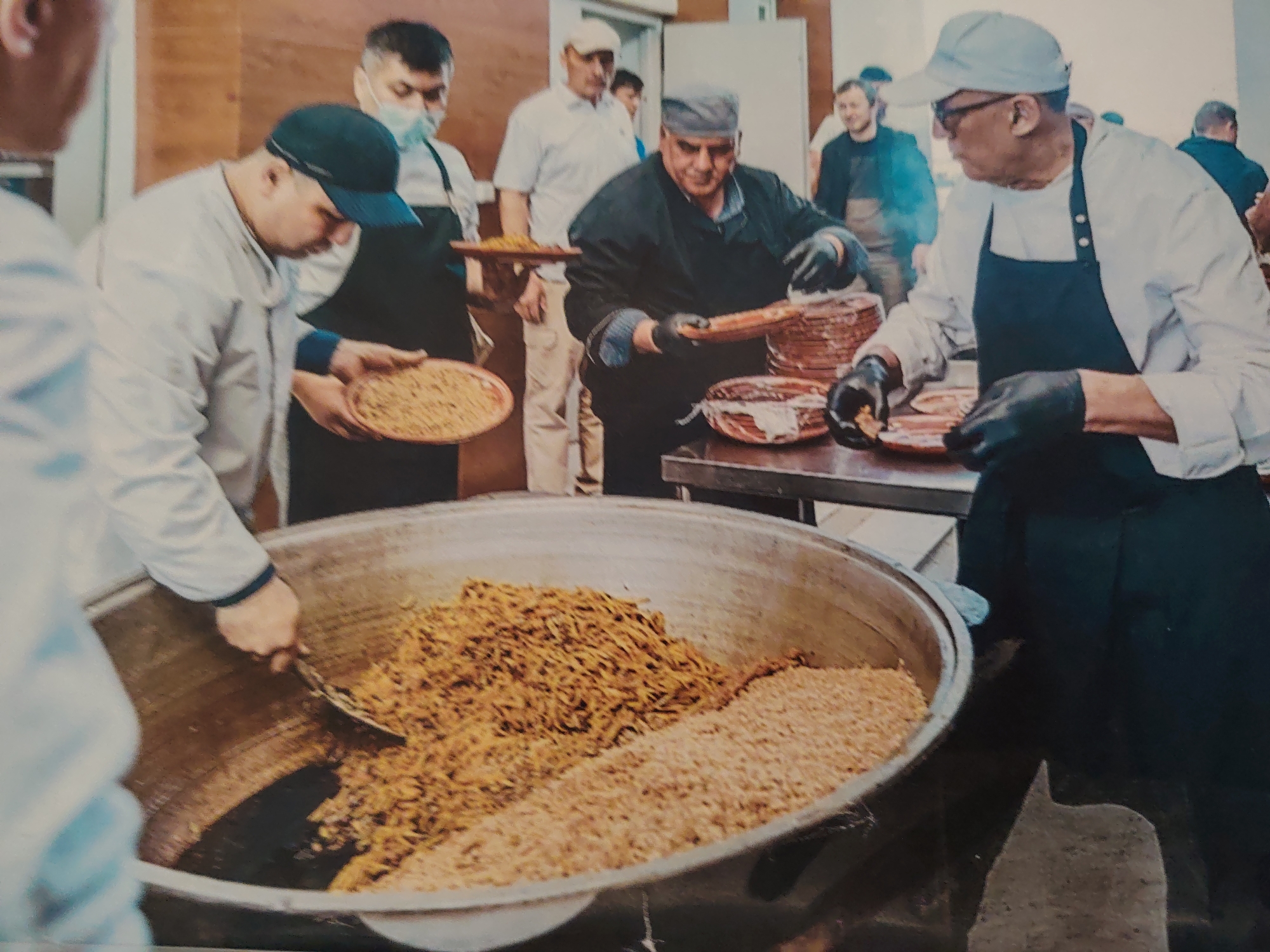 In Konigil, Uzbekistan, a group of men are engaged in food preparation in what appears to be a communal kitchen or event. Dominating the foreground is a large, round metal vat containing two distinct mounds of food: a lighter colored, granular substance, likely rice or grain, and a darker, shredded or chopped material, possibly meat or vegetables. A man in a white coat and a dark blue baseball cap is using a metal scoop to stir the contents of the vat. To the left, another individual, also wearing a white coat and a blue face mask, holds a circular platter filled with a light brown, granular food. In the mid-ground, a man in a dark jacket and a flat cap is working with stacked circular dishes, each appearing to contain a similar light brown granular food. He is wearing black gloves and appears to be arranging or serving the food. Other men are visible in the background, some wearing aprons and head coverings, suggesting a structured preparation process. The environment seems to be indoors with wooden paneling on the walls. The overall scene depicts a large-scale food service operation, possibly for a community event or a large gathering.