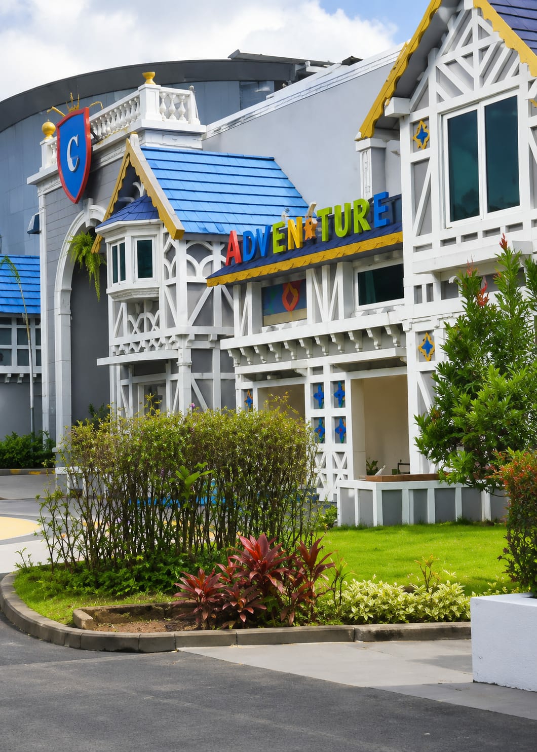The image depicts the exterior of a building with a whimsical, colorful design, likely a theme park or entertainment venue, located in Bandar Baru, Indonesia. The building features a bright blue roof with yellow trim and white decorative elements resembling stylized half-timbering. A sign with the word "ADVENTURE" in multi-colored, three-dimensional letters is prominently displayed above an entrance. To the left, a large archway is visible with a coat of arms featuring a blue shield with a white "C" against a red background. The surrounding landscape includes well-maintained green grass, lush bushes, and some red-leafed plants. In the background, a large, curved, grey structure suggests a more modern adjacent building or stadium. The sky is partly cloudy. There are no people or animals visible in the scene. The lighting suggests it is daytime.