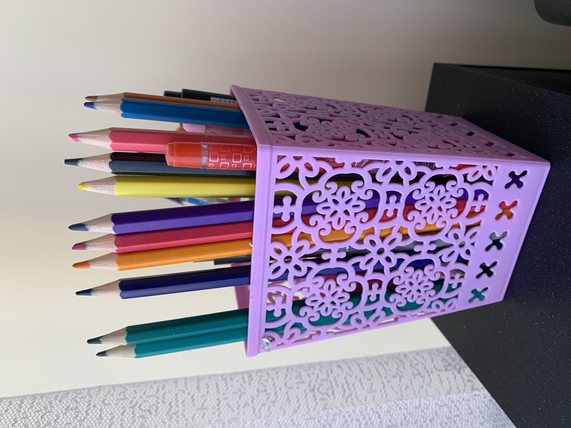 A partially filled, light purple, laser-cut plastic pencil holder sits on a surface, with pencils of varying colors extending out of the open end of the holder. The pencils are arranged roughly horizontally. The background is a white wall on the left, and a dark surface on the right. The scene's location is not discernible from the image.