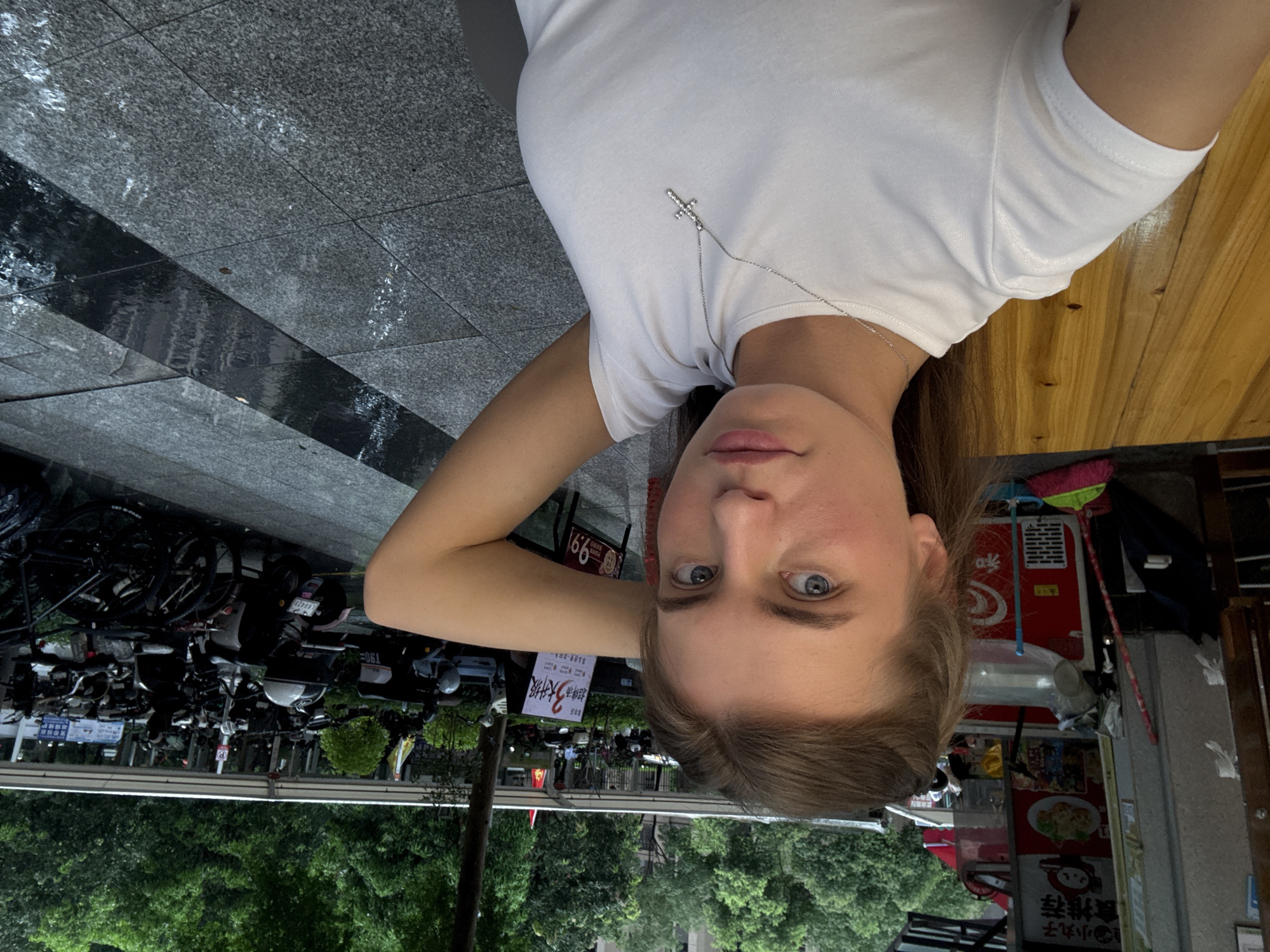 The image shows a young woman with light brown hair, wearing a white t-shirt and a silver cross necklace. She is looking at the camera, with her right arm raised behind her head. The woman is sitting at a wooden table, with a food stall and a row of parked motorcycles and bicycles in the background. The scene appears to be outdoors, with trees and buildings visible in the distance. The ground is paved with stone tiles, and there are puddles of water, suggesting it has recently rained. The food stall has Chinese characters and images of food. The parked vehicles are typical of those found in urban areas of China, specifically in Haizhu District, Guangzhou, China.
