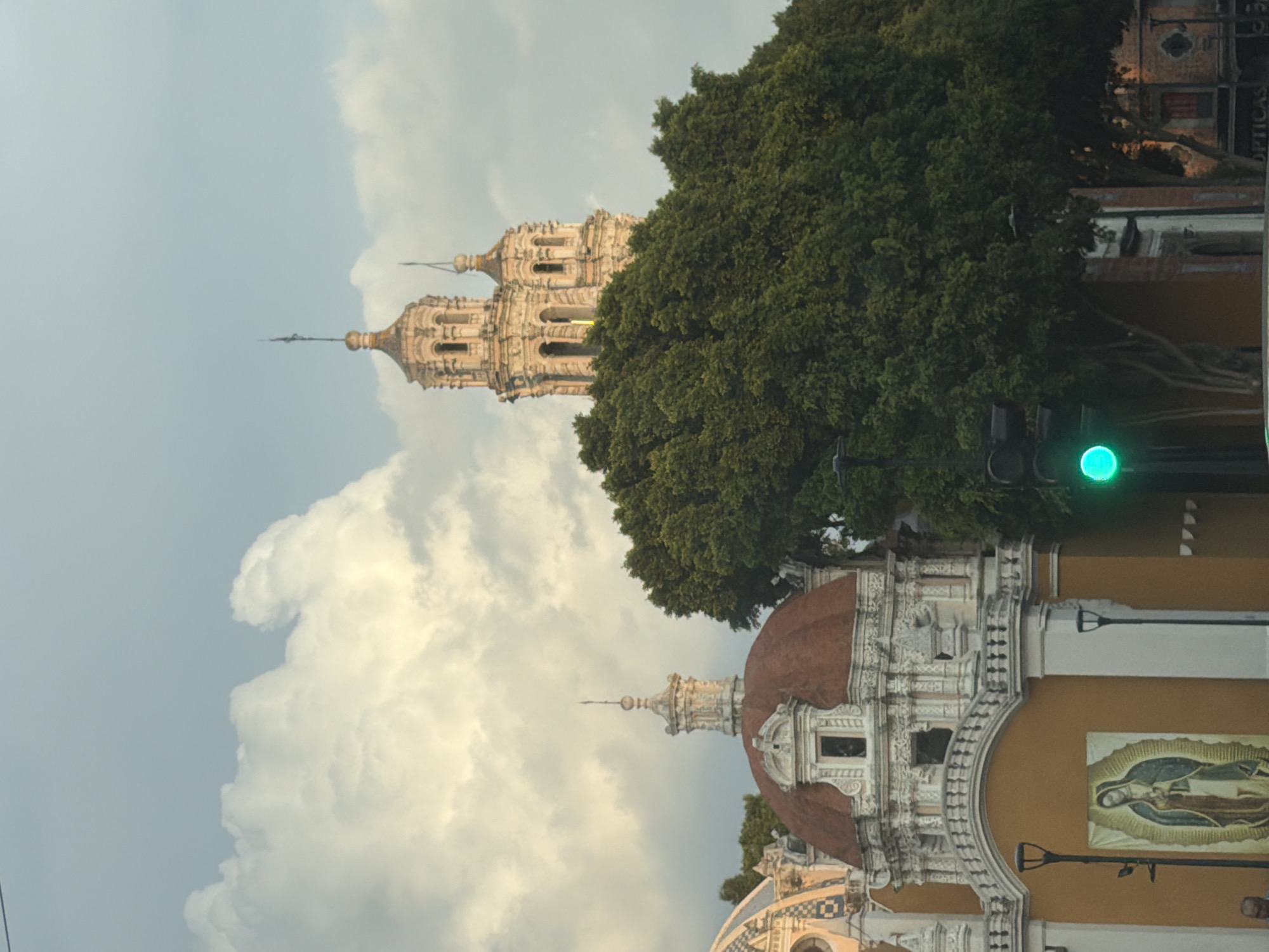 The image shows two historic buildings in Puebla, Mexico. In the foreground is a building with a brown dome, white decorative trim, and a yellow wall displaying an image of the Virgin of Guadalupe. A street lamp is affixed to the wall. A green traffic light is also visible. Behind this structure, partially obscured by a large green tree, stands a multi-tiered tower. The sky is filled with large, white, puffy clouds.