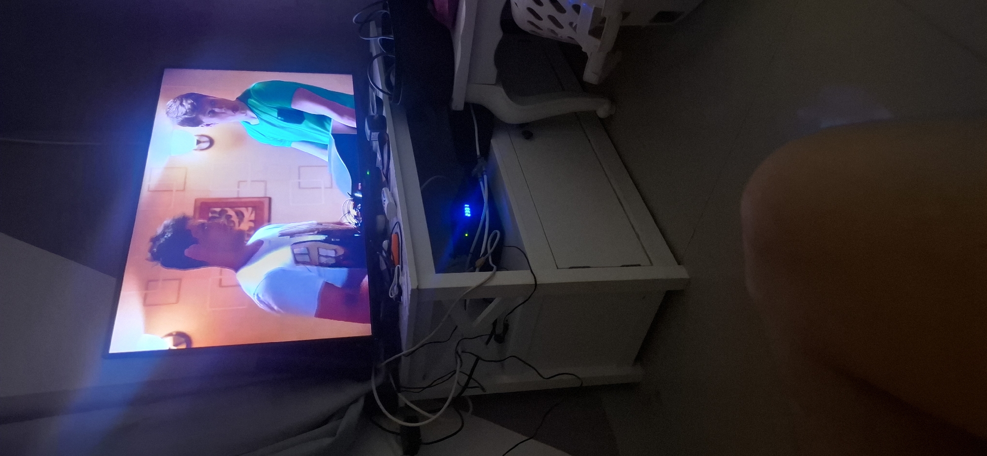 A television screen displays two young men. The man on the left wears a white t-shirt with a graphic. The man on the right wears a green t-shirt. They stand in front of a wall with a decorative pattern and a framed picture. Below the television, a white media console is visible. It is cluttered with various electronic devices and cables. A blue digital display on one of the devices reads "2301". The scene is dimly lit, with a soft glow emanating from the television and the blue display. The floor is a dark, tiled surface. The image appears to have been taken indoors, possibly in a living room, in Yerocham, Israel.