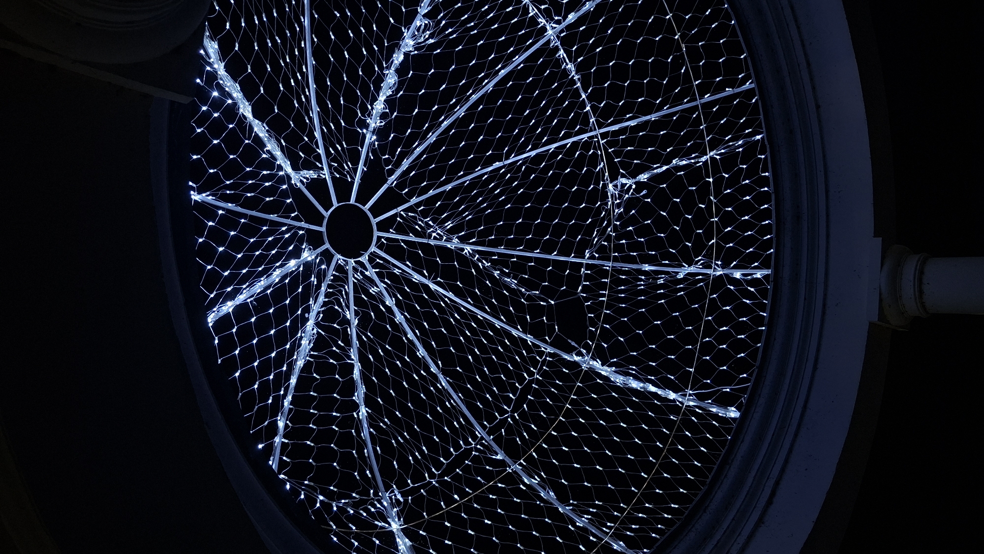 The image shows a geodesic dome structure covered in white LED lights. The dome is viewed from below, looking upwards towards the apex. The frame of the dome consists of radiating spokes and concentric rings. The lights are arranged in a net-like pattern across the dome's surface, creating a glowing, star-like effect against the dark background. In the lower portion of the image, a curved architectural element, possibly a cornice, is visible, along with a partial view of a white column. The scene appears to be an outdoor installation, possibly a festive decoration. There are no people or animals present in the image. The location is identified as Myadzyel, Belarus.