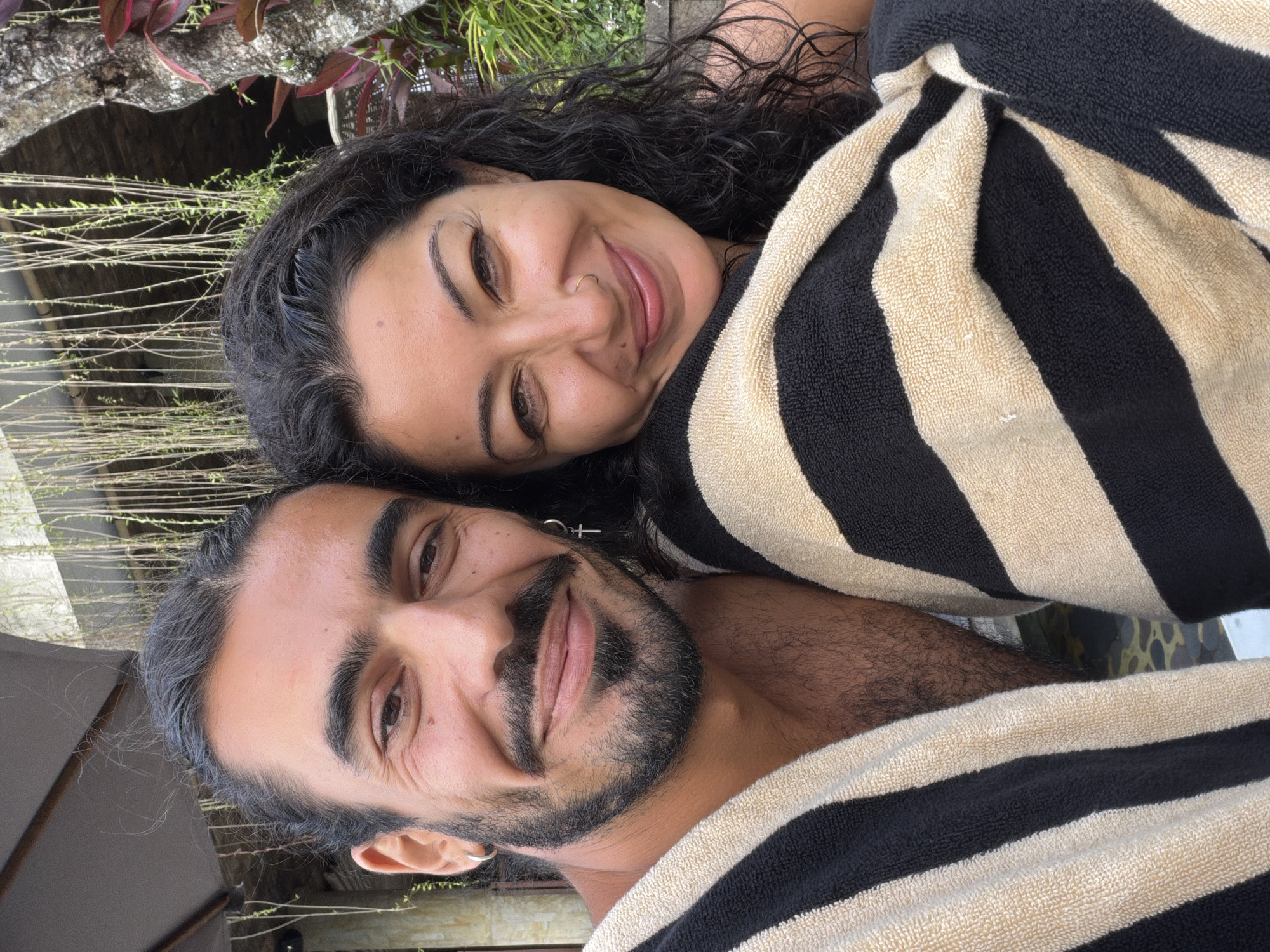 A selfie shows a man and a woman wrapped in striped towels, posing closely together. The man, on the left, has dark hair pulled back, a beard, and a mustache. He wears a hoop earring and a cross earring. The woman, on the right, has dark, curly hair and a nose ring. They are outdoors, with lush green foliage and a tree trunk visible in the background. The environment suggests a tropical setting, possibly a resort or private villa. The image appears to be taken in Tegallalang, Indonesia, known for its rice terraces and scenic landscapes.