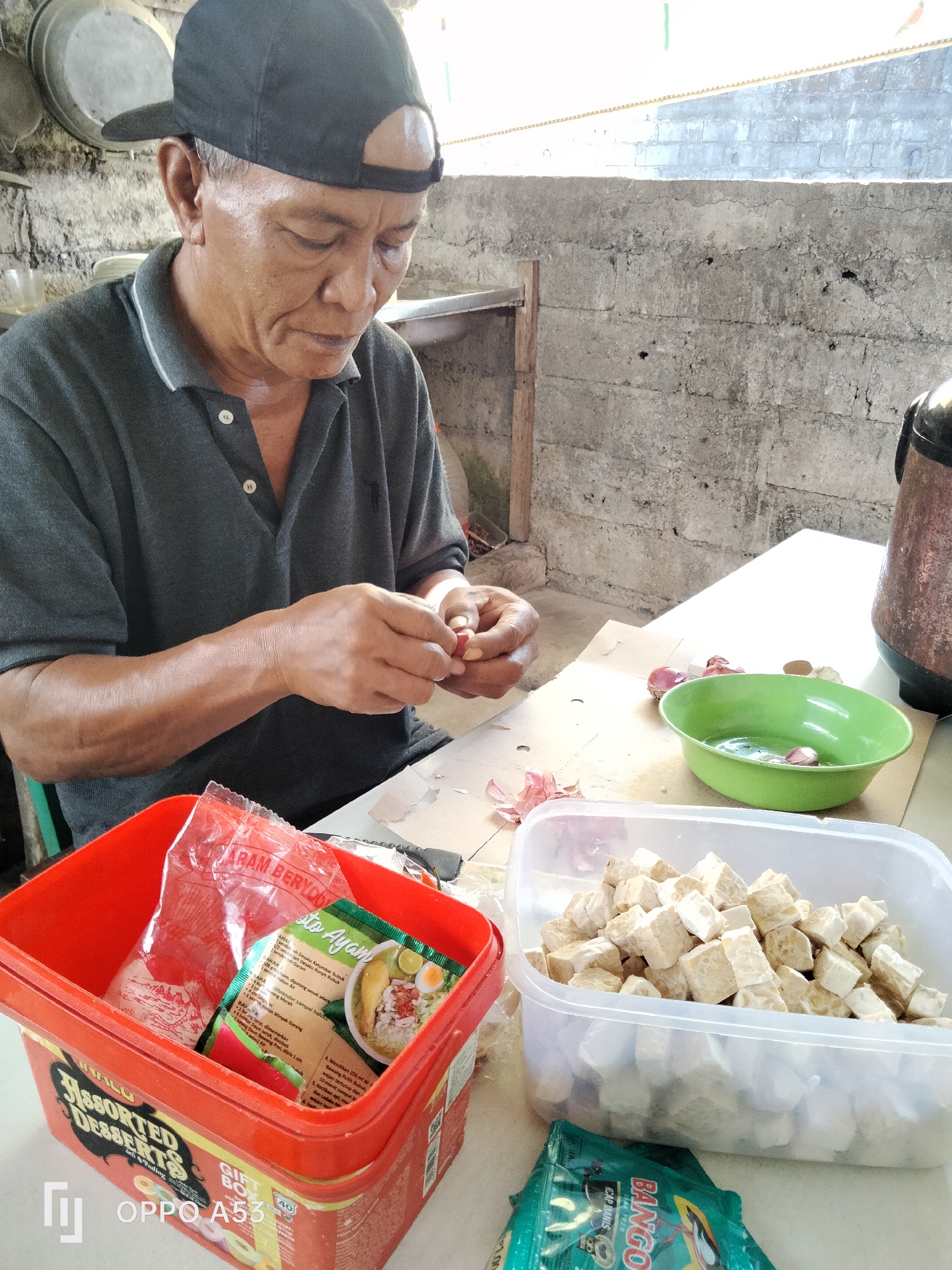 An elderly Indonesian man wearing a black baseball cap and a dark grey polo shirt is peeling garlic over a light-colored, possibly melamine, table in what appears to be an outdoor or semi-outdoor kitchen area. The background consists of a rough, grey concrete block wall, and a portion of a metal or ceramic pot is visible on the right. In front of the man, a red plastic container holds a plastic bag with what looks like seasoning packets and a product label. To the right of the red container is a clear plastic tub filled with diced tempeh. A bright green bowl containing red onions and some liquid is also on the table, positioned near the man's hands. Several peeled garlic cloves and onion skins are scattered on the table. The overall impression is of a domestic scene, likely in a humble setting, where food preparation is underway.