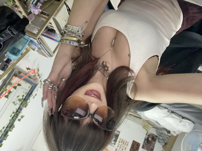 A woman poses for a selfie, wearing a white tank top and large brown sunglasses, with her tongue slightly visible. Multiple rings adorn her fingers, and a collection of bracelets jingles on her wrist. The background includes a cluttered shelf, a mirror with lights, and a glimpse of a room with various items.