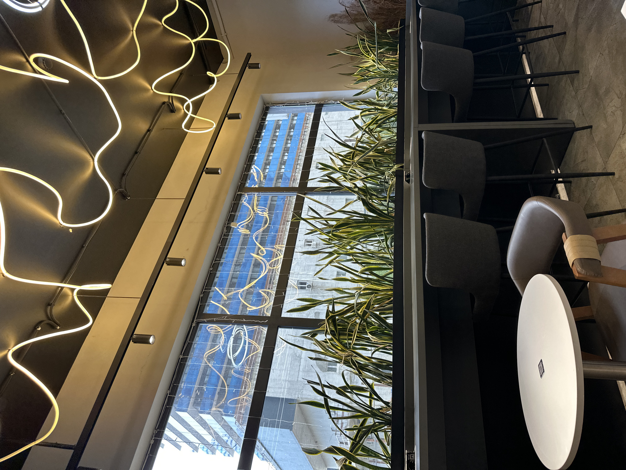 The interior of a modern-styled space, possibly a cafe or restaurant, features a high, angled window offering a cityscape view, likely in Batumi, Georgia, given the architectural style. A large, whimsical neon light fixture is mounted on the ceiling, adding a contemporary design element to the space. Tall, slender plants line the window, creating a natural barrier. A row of dark-colored chairs and a small, round table with a white top suggest a seating area. The overall atmosphere is modern and stylish, with a mix of natural and artificial elements.