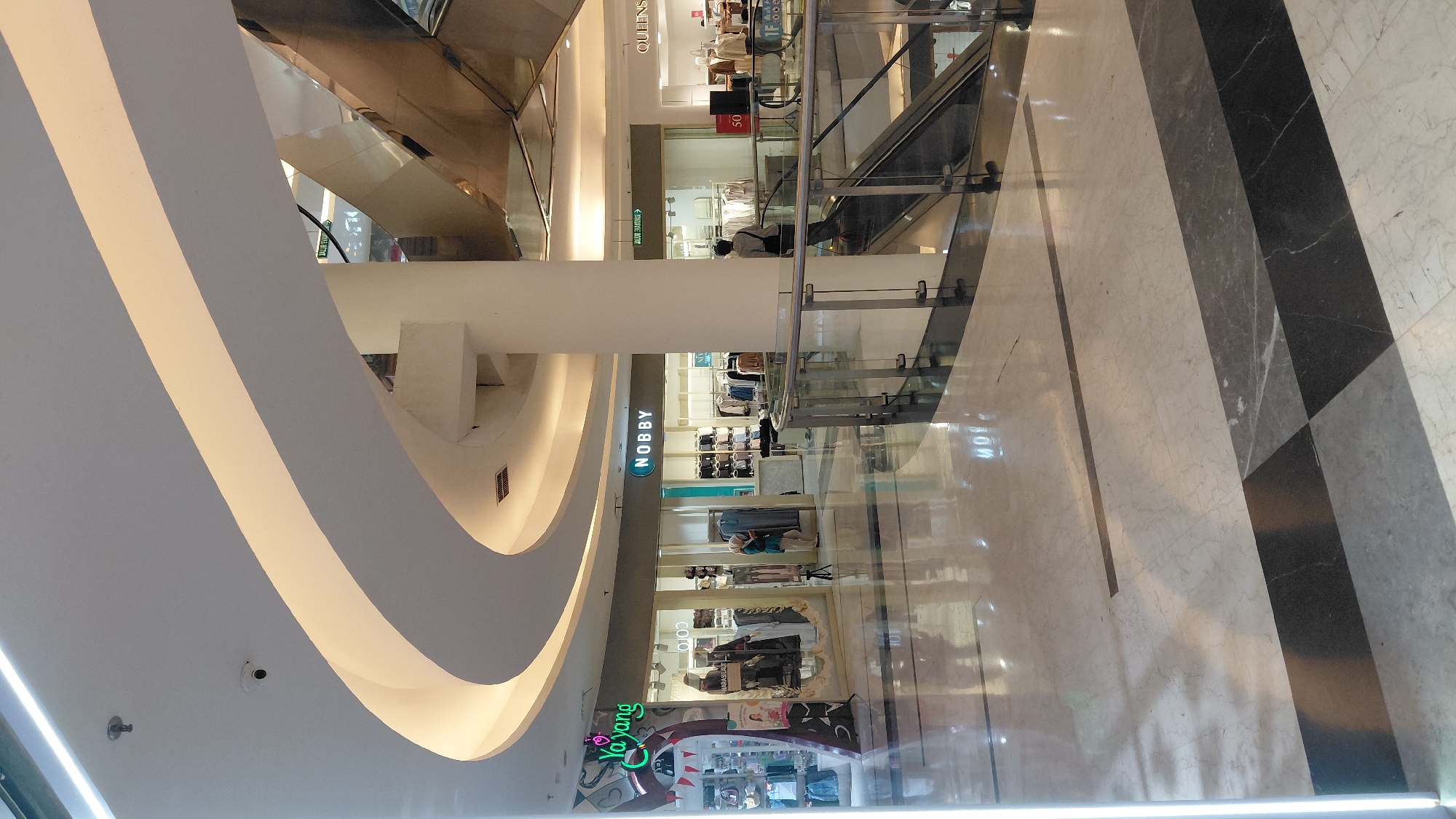 The image captures the interior of a modern shopping mall in Purwokerto, Indonesia. The scene is characterized by a high, curved ceiling with integrated ambient lighting, casting a warm glow on the polished marble floor. In the foreground, the floor displays a geometric pattern of white, grey, and black marble tiles. A clear glass railing with metal supports delineates a walkway and an escalator.
Several retail storefronts are visible. To the left, a store named "YAYANG" is identifiable by its neon green signage. Next to it, another store displays mannequins dressed in clothing. Further to the right, a store with the "NOBBY" logo is seen, with racks of apparel inside. Beyond that, a store with "QUEENS" signage is partially visible. An escalator, clad in stainless steel, ascends towards the upper levels of the mall.
Two individuals are present in the scene. One person, wearing a blue hijab and light-colored clothing, stands inside the "NOBBY" store, facing towards the racks of clothes. Another individual, dressed in a grey top and dark pants, walks away from the viewer towards the escalator. A security camera is mounted on the ceiling near the curved architectural element. The overall impression is of a well-lit, contemporary commercial space.