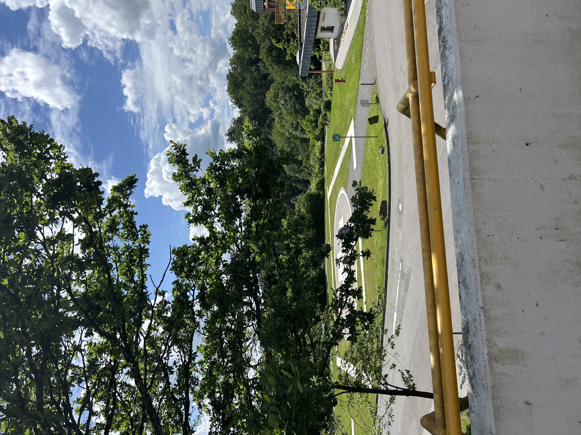 The image captures an outdoor scene in Gummersbach, Germany, viewed from an elevated perspective on a bright, partly cloudy day. In the foreground, a weathered grey concrete wall topped by two parallel yellow metal pipes, serving as a railing, extends across the bottom. A large deciduous tree with dense green foliage partially obscures the upper left and central portions of the scene, its leaves silhouetted against the sky in places.
Below the railing, an asphalt road or path, marked with a white line, transitions to a manicured green lawn. On this lawn, a prominent white 'H' encircled by a white line clearly marks a helipad, with an additional white guiding line extending from it. Adjacent to the helipad are a small green sign and a brown informational sign, likely bearing German text, possibly "Flugplatz." A red fire hydrant is visible on the grass near a building.
To the right, a modern multi-story building, possibly part of a complex, features large windows and a flat roof. A large yellow letter 'K' is visible on its exterior, and the word "Mediana" is partially legible near an entrance, strongly indicating a hospital or medical facility, such as the Mediana Klinikum Gummersbach. The background consists of a dense, verdant forest or wooded area, suggesting a natural setting surrounding the facility. The sky is bright blue with scattered white cumulus clouds. No people are visible, and no specific actions are occurring in the static scene.