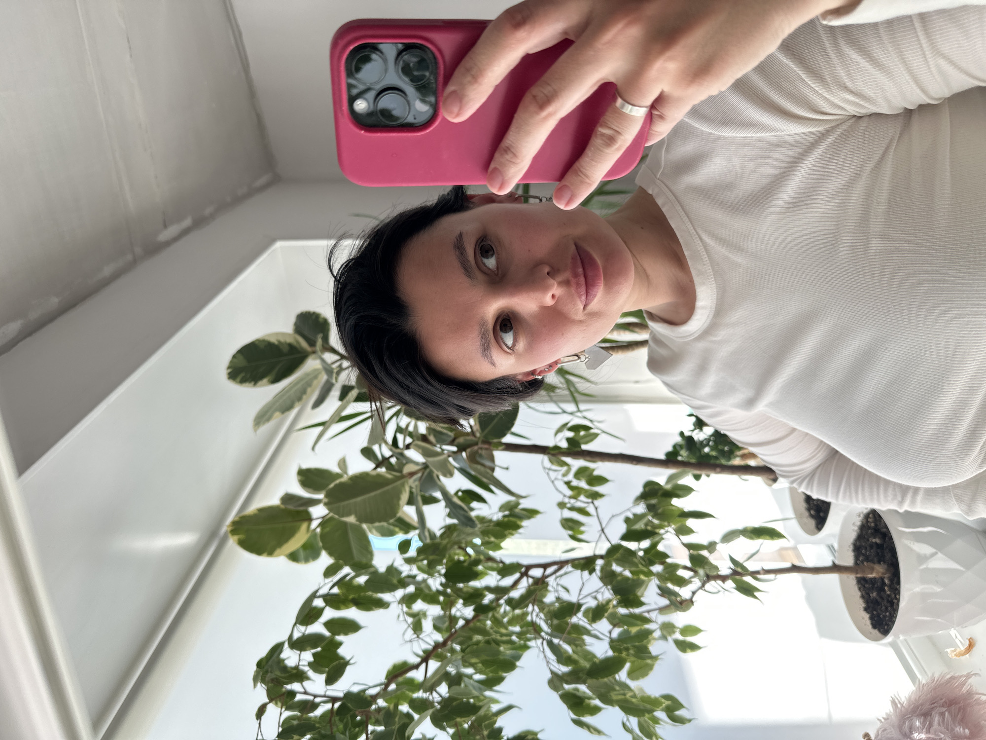 A woman is taking a selfie indoors. She is wearing a white shirt and holding a pink phone. She has short dark hair and is wearing earrings and a ring. Behind her is a window with several potted plants. The location is suggested to be in the city of Chaikovsky, Russia.