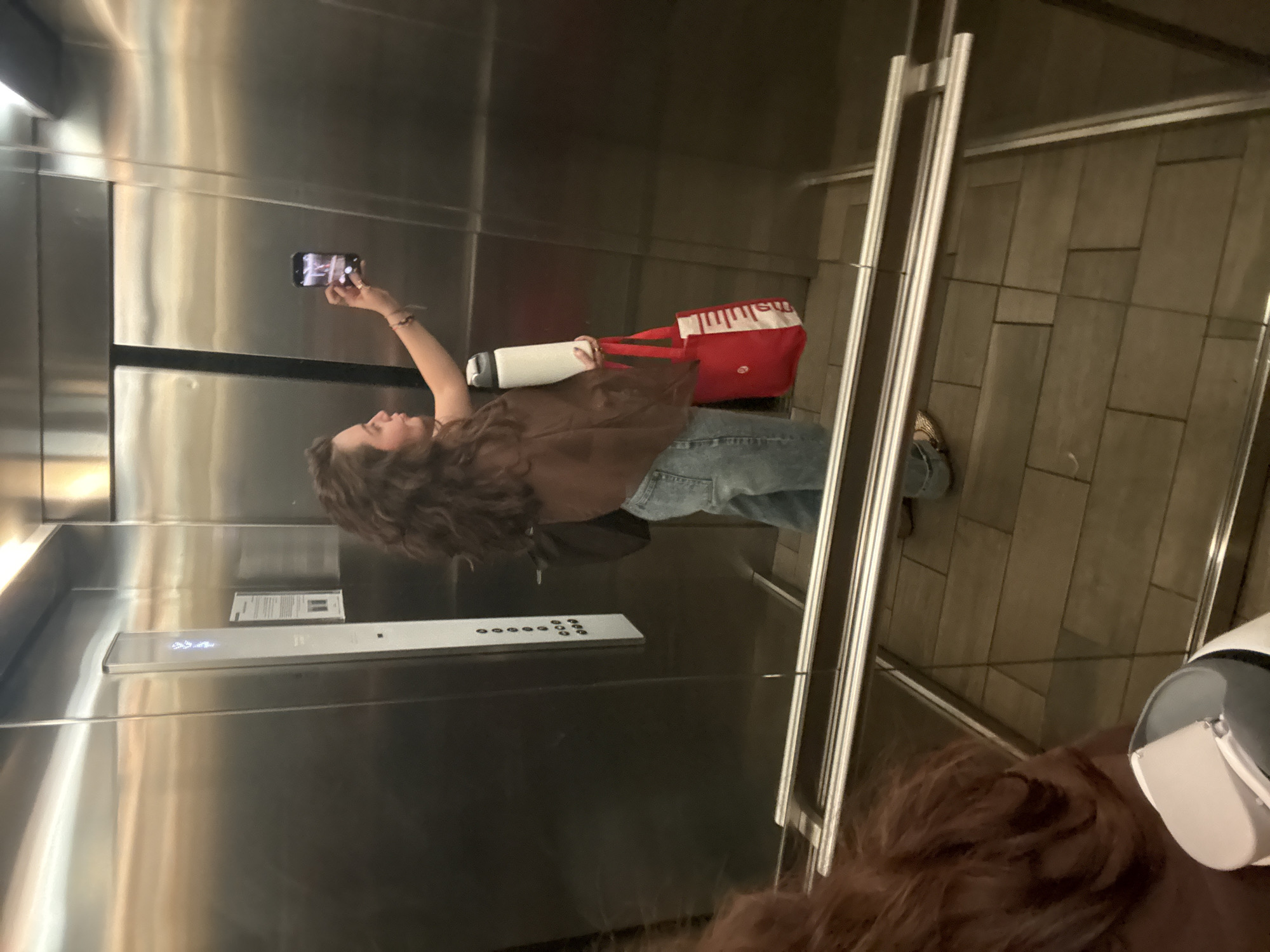 A woman with curly brown hair is taking a selfie in an elevator. She is wearing a brown jacket and blue jeans. She is holding a cell phone in her right hand. She is also holding a water bottle and a red tote bag. The elevator has a stainless steel interior. The floor of the elevator is tiled. The current level is showing on the display. The location is most likely somewhere in Mexico City, Mexico.