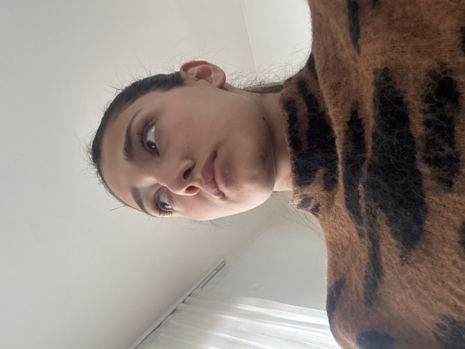 A medium-close-up, low-angle shot features an adult female, visible from the chest upwards, against an indoor background. She has a medium complexion, dark hair pulled back, and is wearing a high-necked, textured sweater with a brown and black animal print pattern. Her head is angled slightly upwards and to the right, with her gaze directed towards the upper left portion of the image frame. Her lips are slightly parted. The background consists of a plain, light-colored ceiling and wall. To the left, a window covered by translucent white sheer curtains is visible, along with a partial view of a white horizontal vent above the window frame. The image provides no discernable visual information to confirm the location as Cibukovac, Serbia.