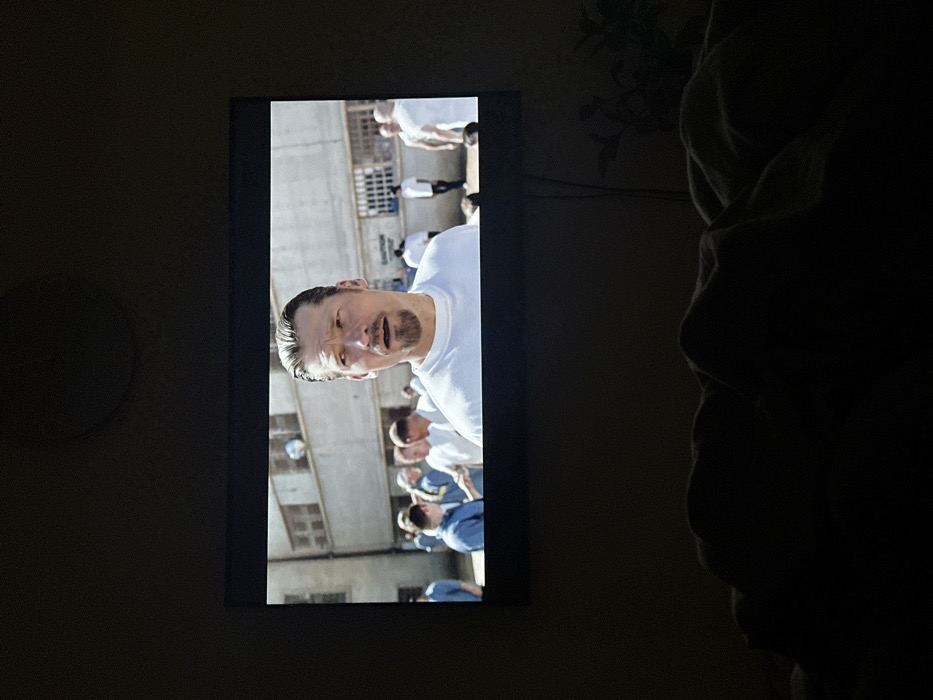 The image depicts a television screen playing a video, situated within a dimly lit room in Kommunarka, Russia. The screen displays a medium shot of a male individual, appearing Caucasian, with short brown hair slicked back and a goatee, wearing a white t-shirt. His mouth is slightly open, conveying a focused or surprised expression. Behind him, multiple other males, some in light blue button-up shirts and others in white t-shirts, are visible in what appears to be an outdoor, sunlit yard. A large, light-colored building with multiple rectangular windows, some barred, forms the background, suggesting an institutional or prison environment. A light-colored spherical object is visible high on the building to the left. The immediate foreground of the image is dark and out of focus, showing the contours of what appear to be blankets or bedding, indicating the viewer's position in bed. To the right of the television, a dark, silhouetted plant is partially visible, along with a vertical cable running from the television area. The television screen is the sole significant light source in the otherwise dark room.