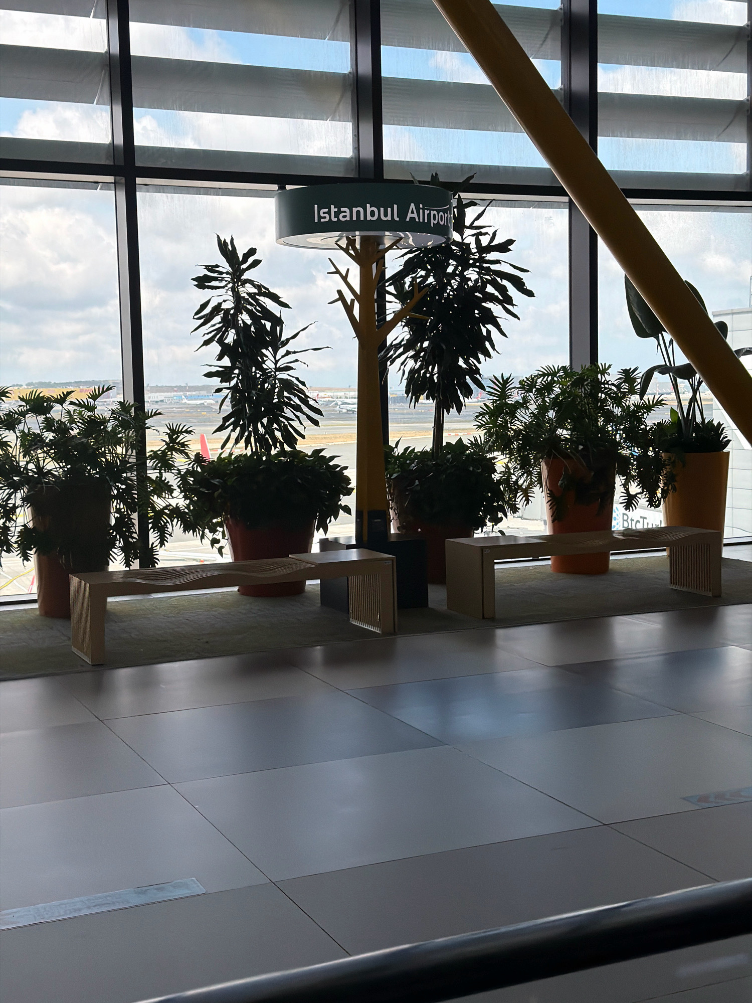 The image shows an interior view of an airport terminal, likely Istanbul Airport, located in Arnavutköy, Turkey. Large windows overlook an airport tarmac with airplanes visible in the distance. Several potted plants are arranged in front of the windows, some in large terracotta-colored planters. A modern, light-colored bench with a curved design is placed in front of the plants. Above the plants, a sign with the text "Istanbul Airport" is visible, attached to a decorative tree-like structure. The floor is tiled in a neutral color with a reflective surface. The overall scene suggests a waiting or lounge area within the airport.