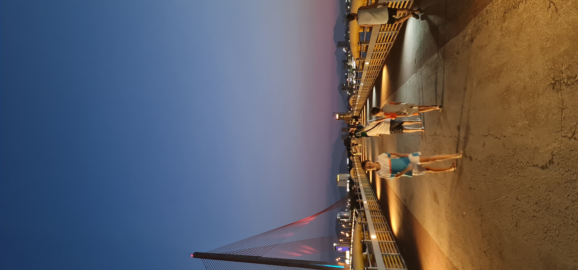 The image depicts an evening scene on a bridge or elevated pathway, likely in a city like Đà Nẵng, Vietnam. The sky displays a beautiful twilight gradient, shifting from a deep blue overhead to a lighter, purplish hue towards the horizon, indicating dusk or early night.
On the right side of the frame, a textured concrete pathway is brightly illuminated by warm yellow lights embedded along a railing. These lights cast long, distinct shadows of the people walking. Several individuals are present on the pathway; in the foreground, a person is seen in a white and blue top with white shorts. Further back, two more people walk, one in an orange-striped shirt and another in a white top and dark shorts, with another individual leaning against the railing further down. All are dressed casually, seemingly enjoying an evening stroll.
On the left, a prominent cable-stayed bridge dominates the midground, its intricate cables and structure outlined by vibrant red lights. In the background, beyond the bridge and railing, the lights of a city twinkle, indicating buildings and urban activity. Distant mountain ranges are silhouetted against the fading sky. The overall atmosphere is calm and pleasant, with the artificial lights creating a warm contrast against the cool twilight sky. There is no visible text in the image.