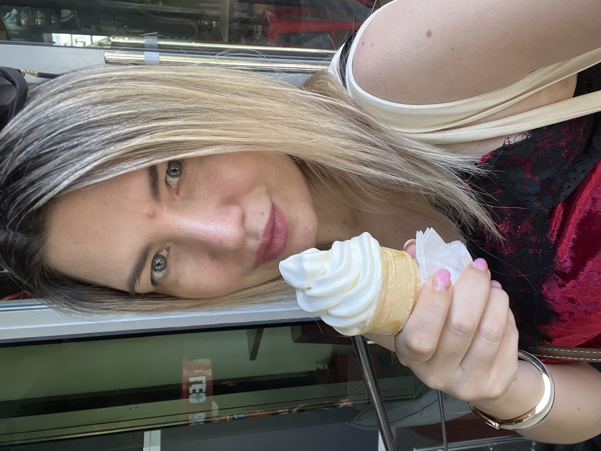 A selfie of a woman with blonde hair and green eyes is shown, holding a vanilla soft-serve ice cream cone in her right hand. She is wearing a red velvet top with black lace trim and a cream-colored strap from a bag. A silver bracelet adorns her left wrist. The background suggests an outdoor setting, possibly near a building with glass windows and reflections of signs that are not clearly legible, though "FEST" is partially visible. The setting is described as being in the city of Sochi, Russia.