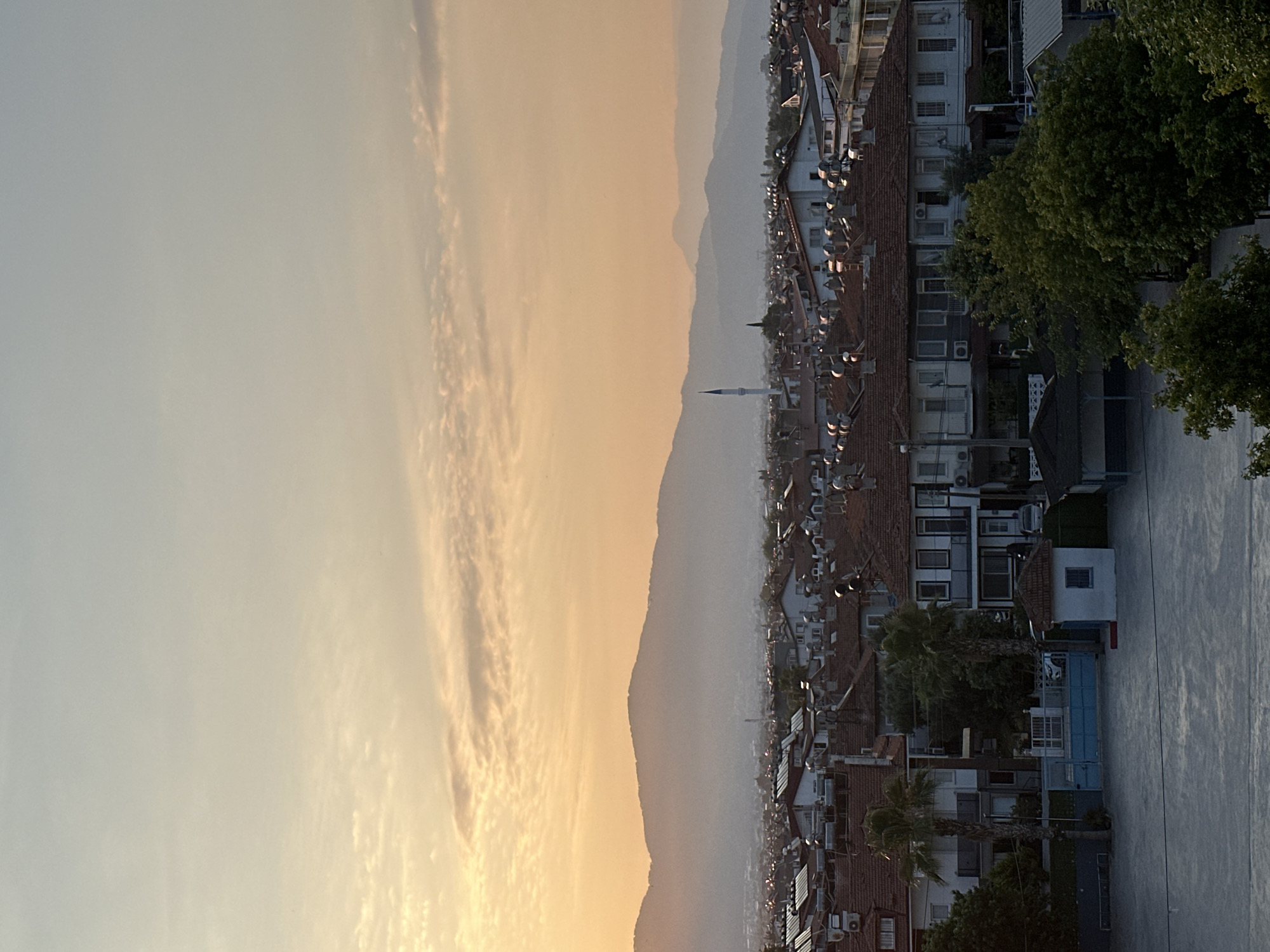 This is a medium-shot, high-angle, outdoor photograph of a residential area in Fethiye, Turkey, during a sunset. The sky is a gradient of pale blue at the top, transitioning to a soft orange and yellow hue near the horizon. Several wispy clouds are visible, illuminated by the setting sun. In the middle ground, a range of hazy, silhouetted mountains forms the backdrop. Below the mountains lies a dense collection of buildings with red-tiled roofs, characteristic of Fethiye's architecture. A slender white minaret of a mosque pierces the skyline amidst the rooftops.
In the foreground, a courtyard or open space paved with concrete or asphalt is visible. To the left of the courtyard, there are some low-lying structures and trees. Further into the scene, the residential buildings are more detailed. They appear to be two or three stories high, with white or light-colored facades. Many of the roofs have solar water heaters. Two palm trees are prominent in front of one of the buildings. A blue-gated entrance with white railings is also present. On the right side of the image, lush green trees and some covered structures are visible. The overall impression is of a peaceful evening over a bustling urban landscape. No people are visible in the frame.