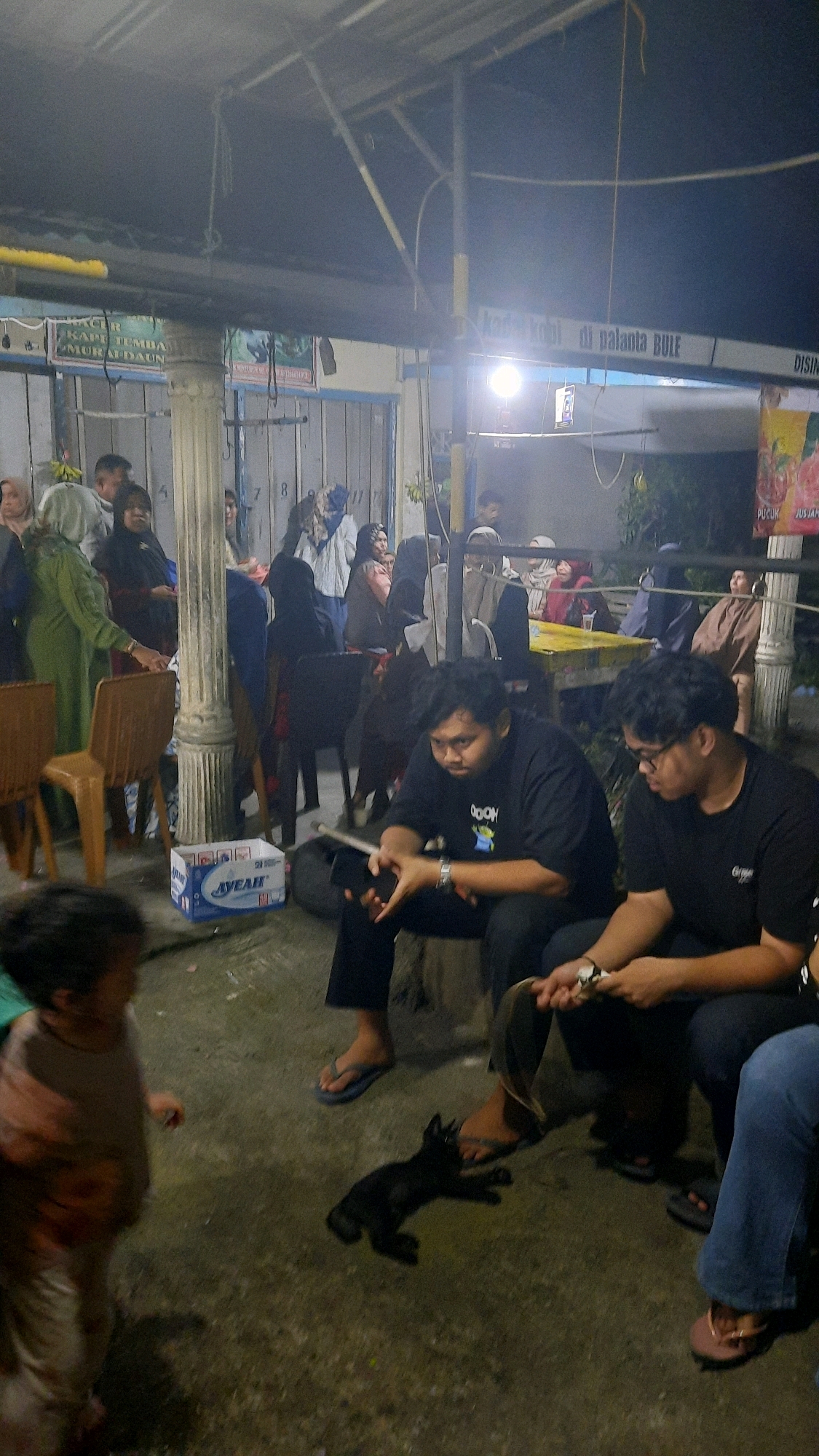 The image depicts an outdoor gathering at night, likely in a community space or an area with covered seating, possibly in Padang, Indonesia. In the foreground, a young child is visible on the left, partially out of focus, appearing to be walking. Next to the child, on the ground, is a small black cat, seemingly lying down.
Further into the scene, two young men are seated. The man on the left wears a black t-shirt with a graphic print and is looking down at his phone. The man on the right wears a black t-shirt and glasses, also looking down at something in his hands. Both men are wearing flip-flops. Their legs and feet are visible.
In the background, a group of people are gathered, some sitting, others standing. They appear to be engaged in conversation or observing something. The environment includes pillars, a roof structure, and signage with text that is partially visible. One sign in the upper right corner has text that is difficult to decipher, but appears to be in Indonesian. There are also chairs and a table visible in the mid-ground. A white box with blue lettering, possibly a product container, is placed on the ground near the pillars. The lighting suggests artificial light sources, typical of an evening event. The overall atmosphere appears to be casual and communal.