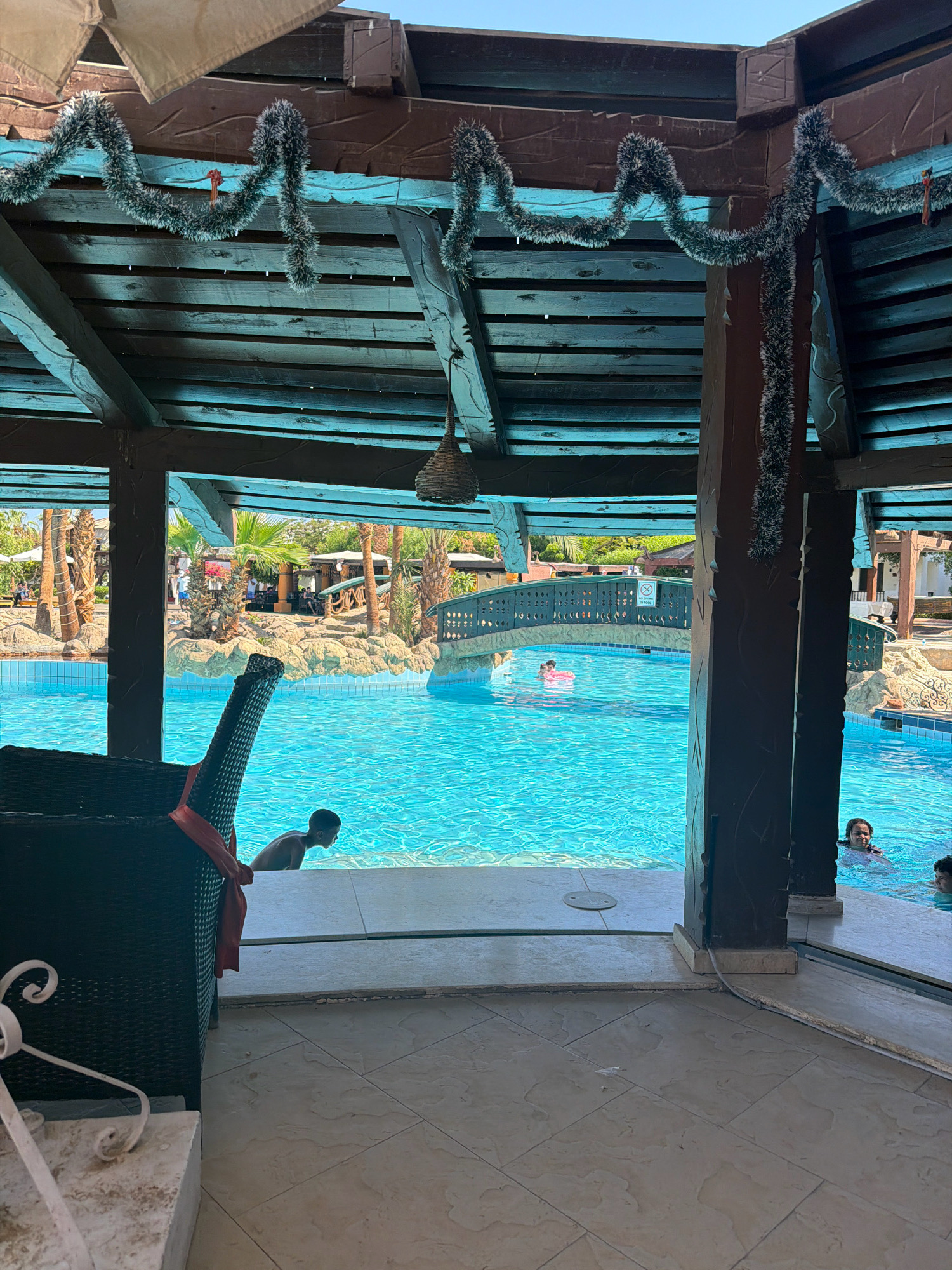 This image shows a view of a swimming pool area at a resort in Sharm El Sheikh, Egypt. In the foreground, a dark-colored wicker chair with a red ribbon tied around its back is positioned on a tiled floor, partially obscuring the view. Beyond the chair, a boy is partially visible at the edge of the turquoise swimming pool, looking out. The pool itself is expansive, with a decorative bridge spanning across it in the midground. Several people are in the pool, including a person in a pink inflatable ring and another person swimming. The background reveals lush green palm trees and other resort buildings, suggesting a tropical vacation setting. The area is covered by a wooden pergola with decorative garland hanging from the beams. The sunlight indicates it is daytime.